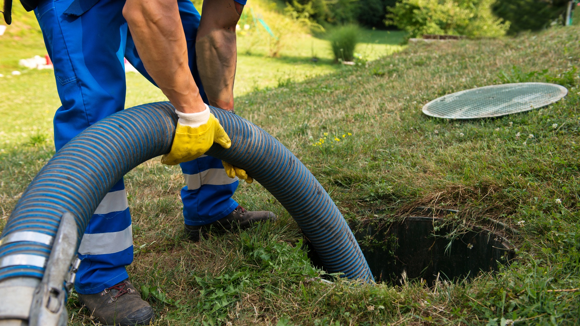 A sewer professional feeding a large hose into the septic tank of a house. The tank is an open hole in the ground.