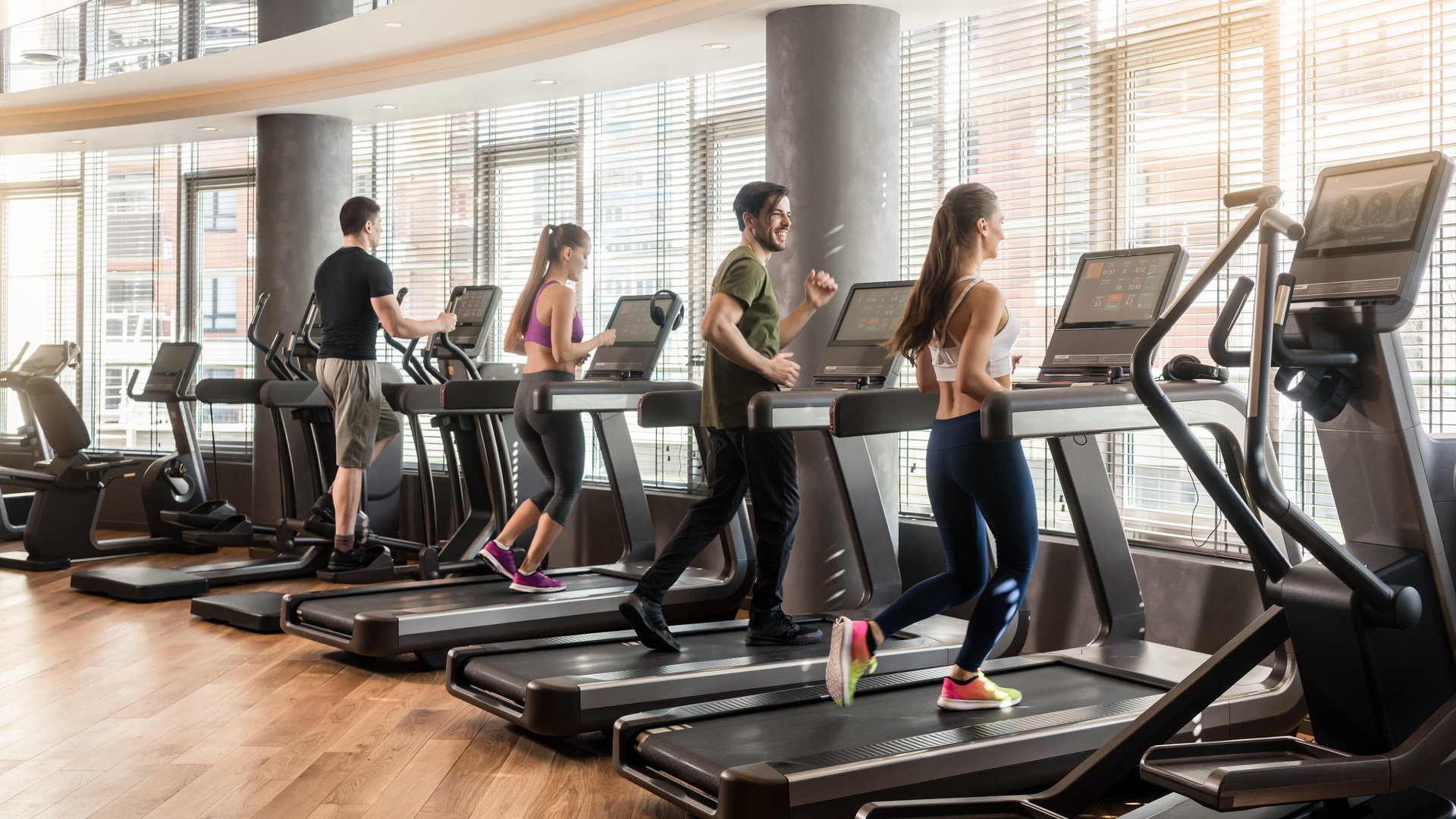 Four people running on treadmills in a bright gym with exercise machines and large windows behind them.