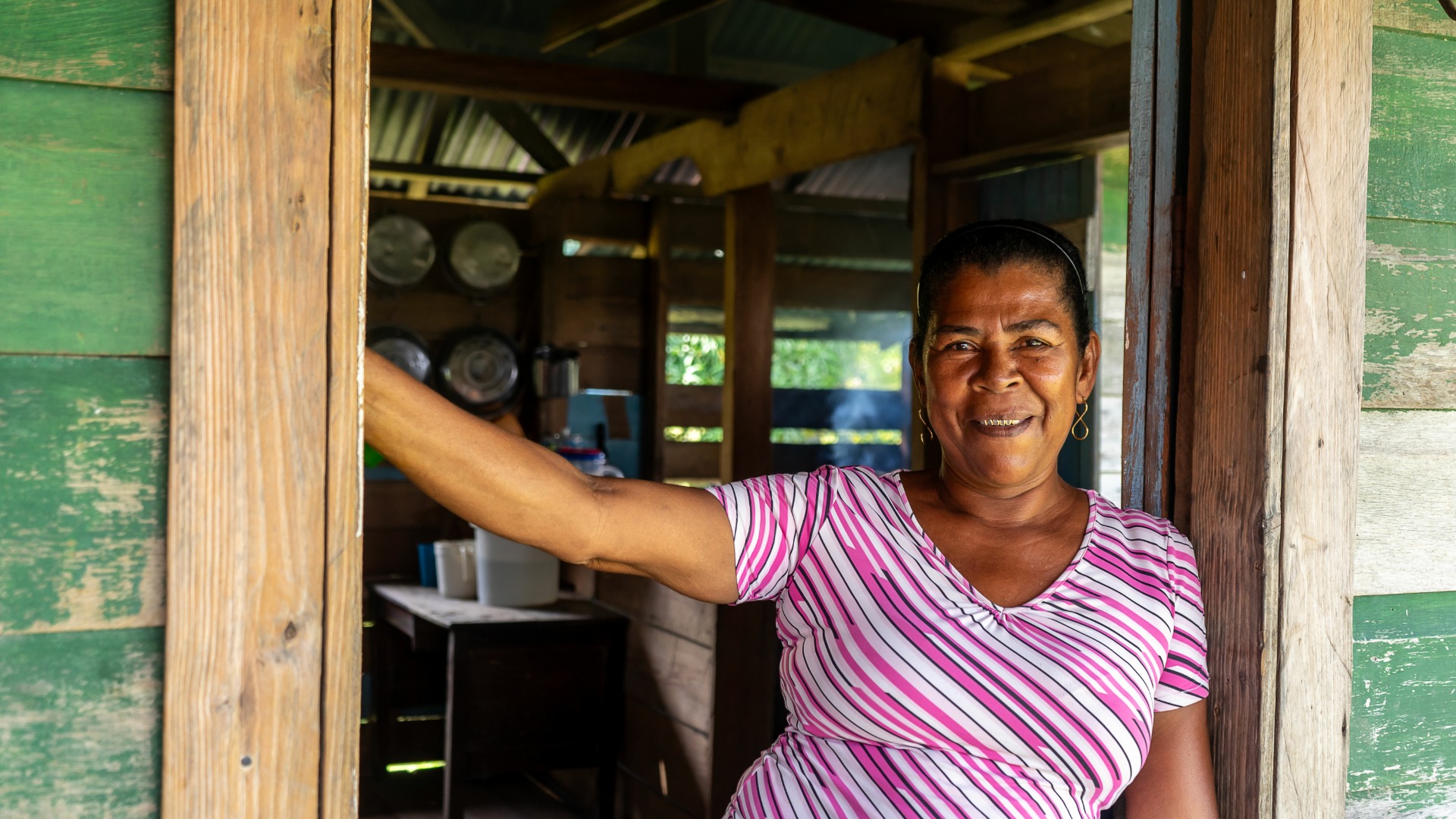 A smiling woman stands in a rustic doorway. Her arm rests on the wooden frame, with kitchen items visible behind her.