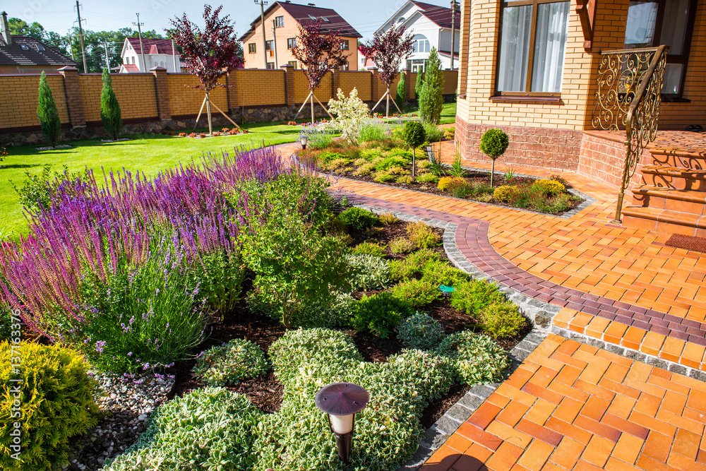 Landscaped garden with purple flowers, trimmed shrubs, green lawn, and curved brick paths beside a brick home.