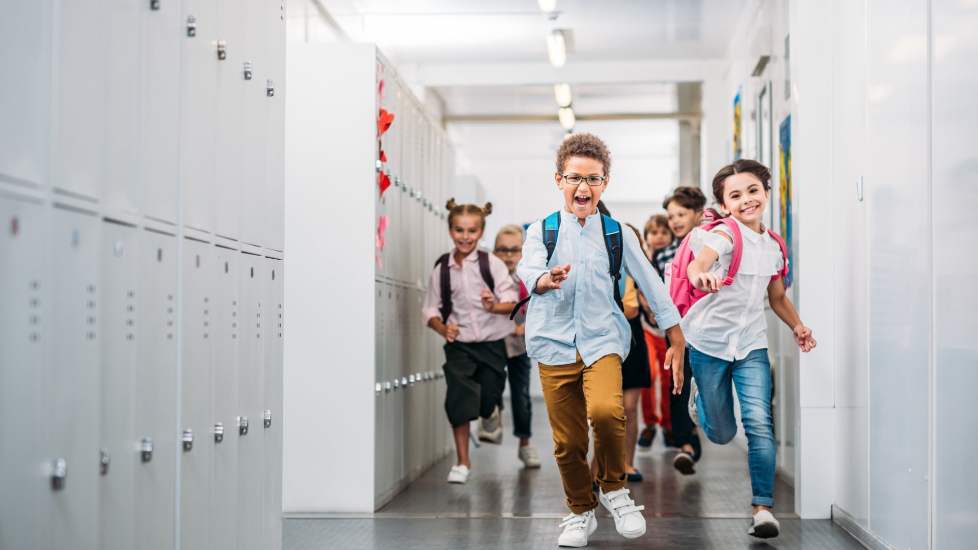 A group of children with backpacks running through a bright school corridor lined with white lockers.