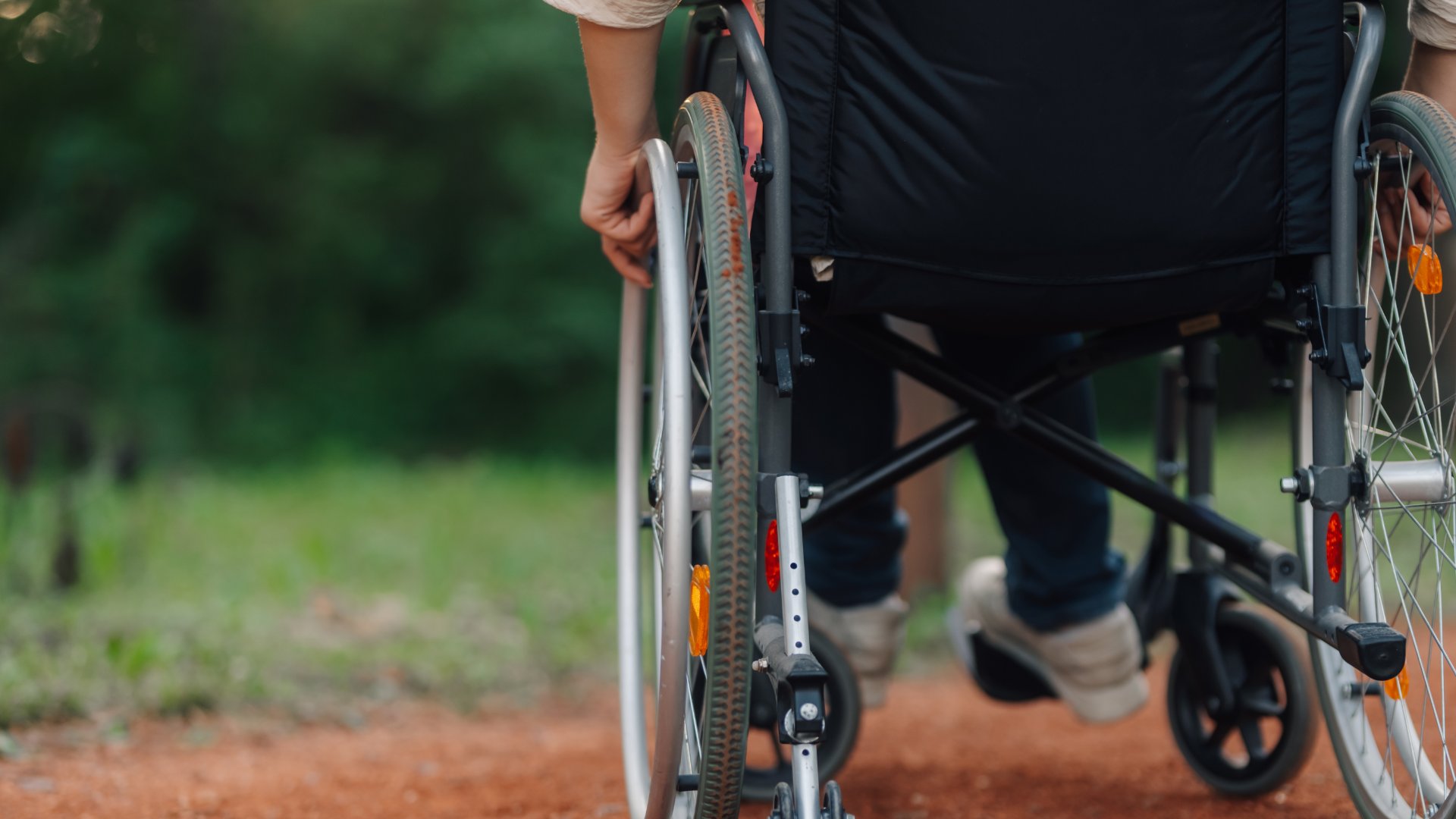 A person in a wheelchair sitting in an outdoor space on a rough path. The person has their hands on the wheels to move.