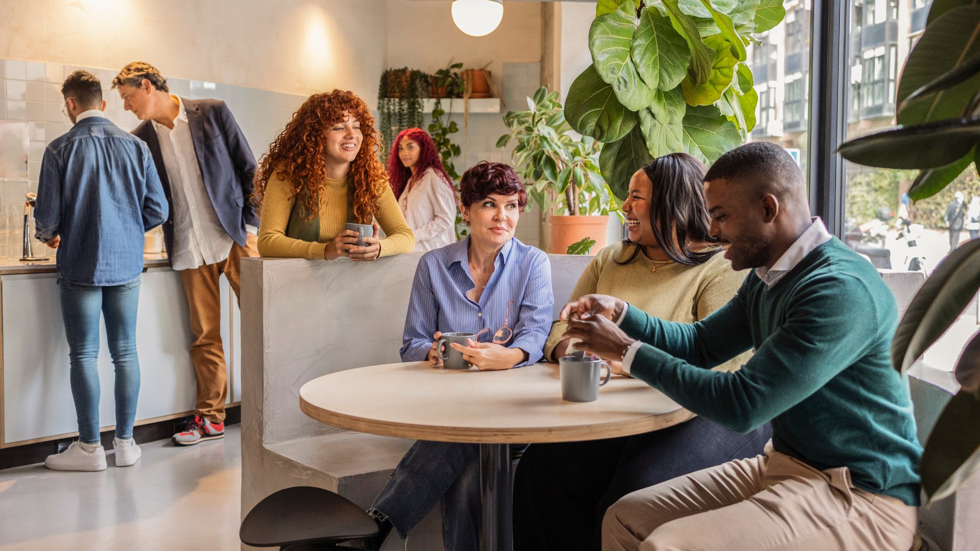 Several colleagues gathered together in an office break room. Some are standing at a counter while others are around a table.