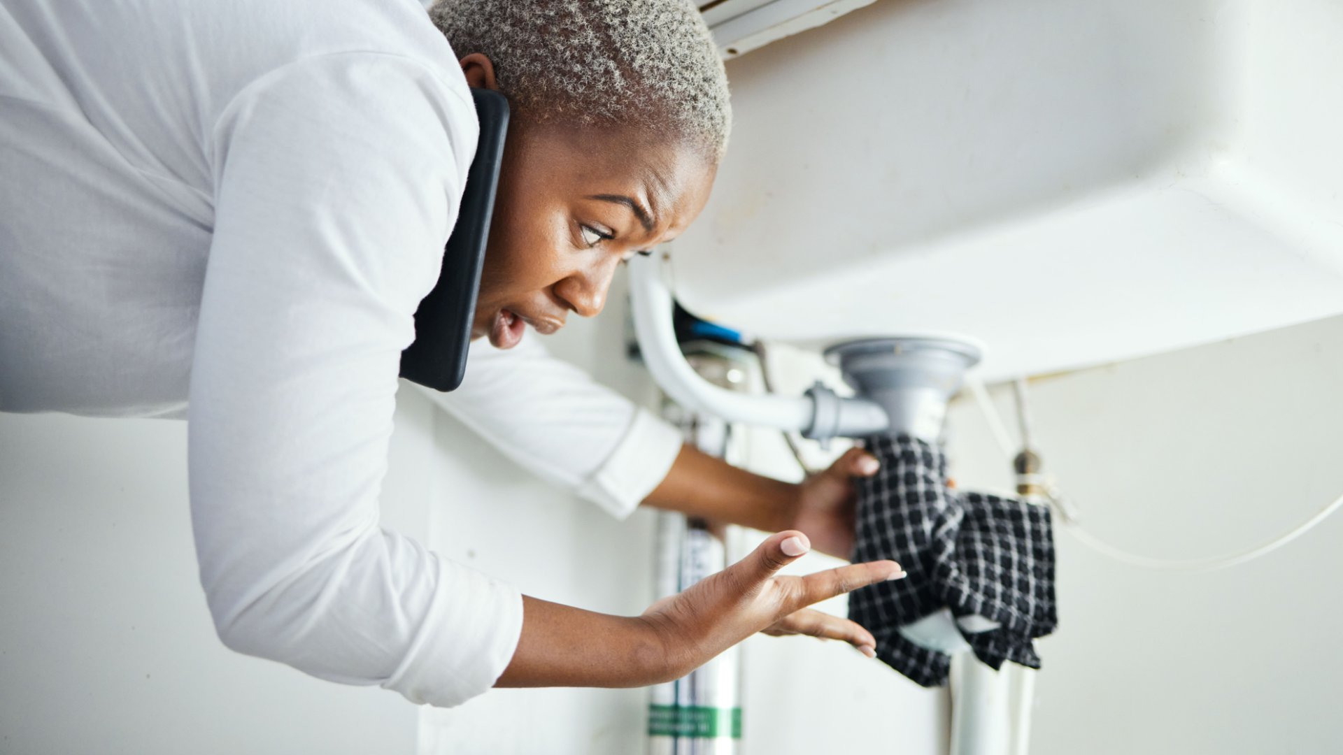 A woman leaning under a leaking faucet. She is on the phone while holding a cloth to the pipe to reduce the leak.