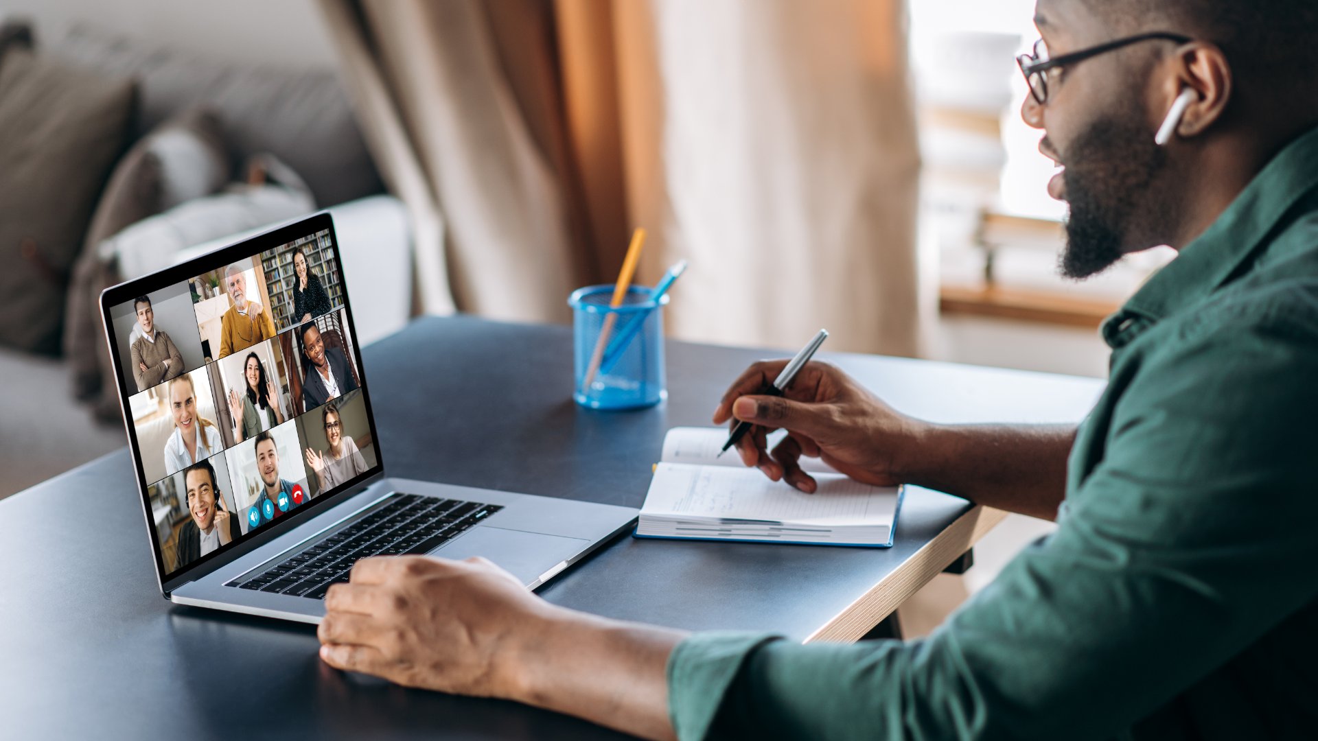 Man wearing earbuds takes notes during a video conference with several coworkers on his laptop at a home desk.