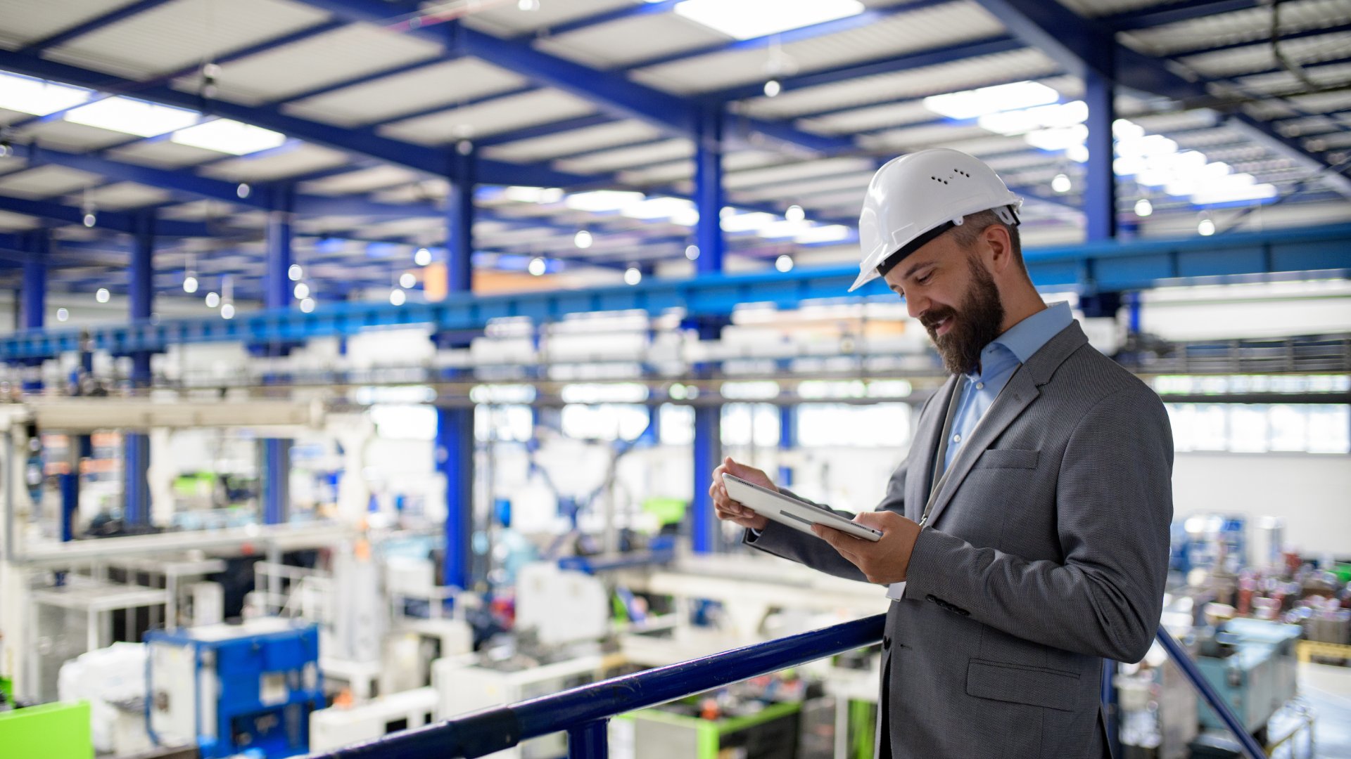 A man in a suit and hard hat using a tablet while overlooking machinery inside a large industrial facility.