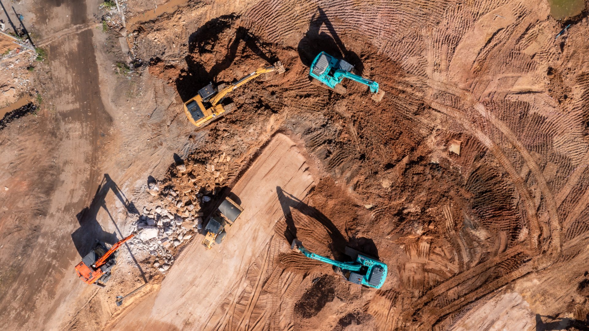 A dirt construction site with several excavators positioned around piles of earth and deep track marks across the ground.