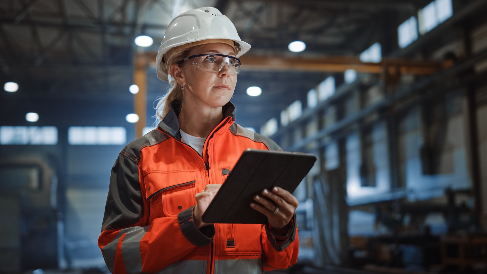 A manufacturing worker in a warehouse is looking at something while holding a tablet. She is wearing safety gear.