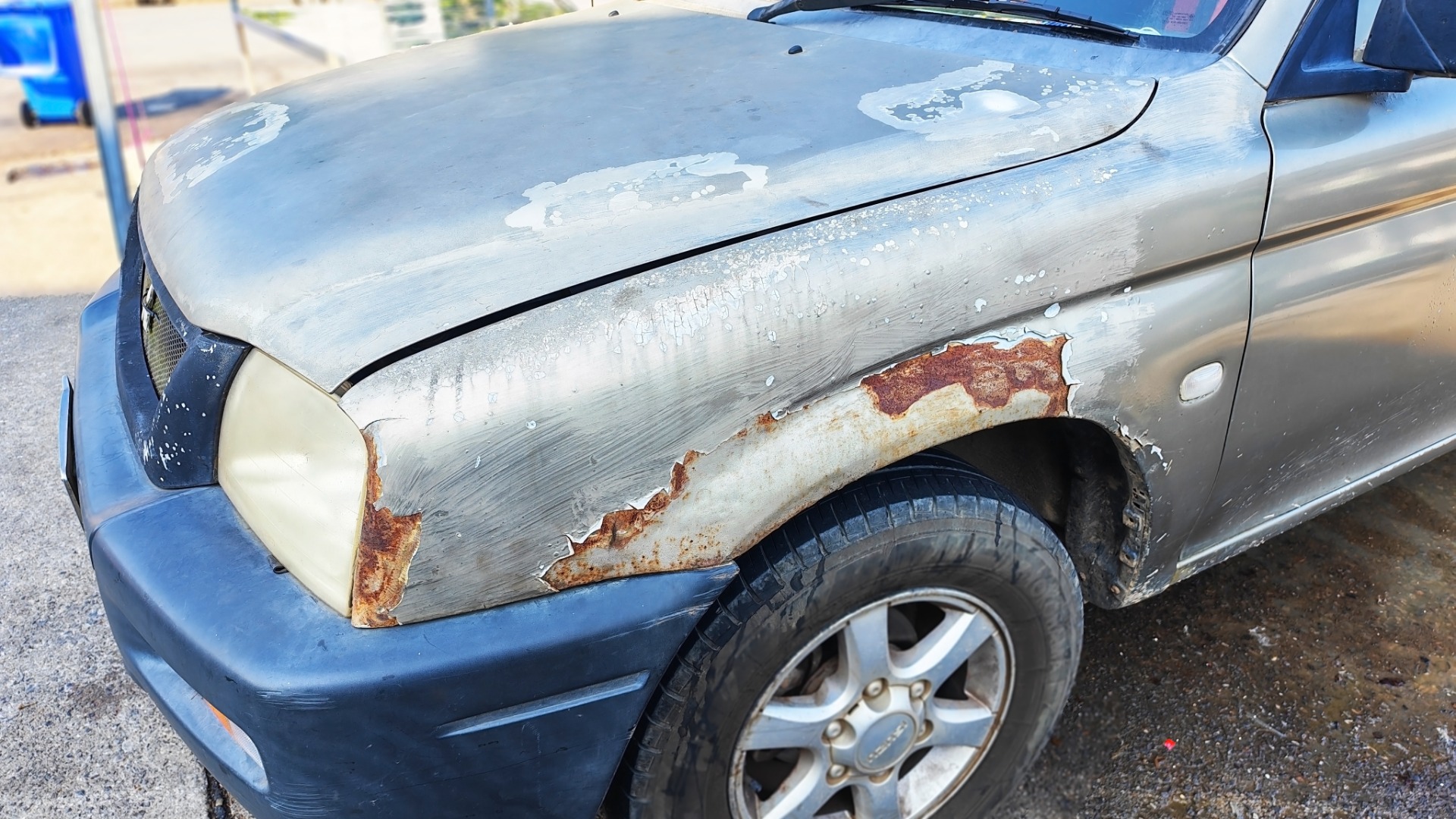 An older vehicle with various damage. There is faded and rusted paint across the hood, while the front bumper is darker.