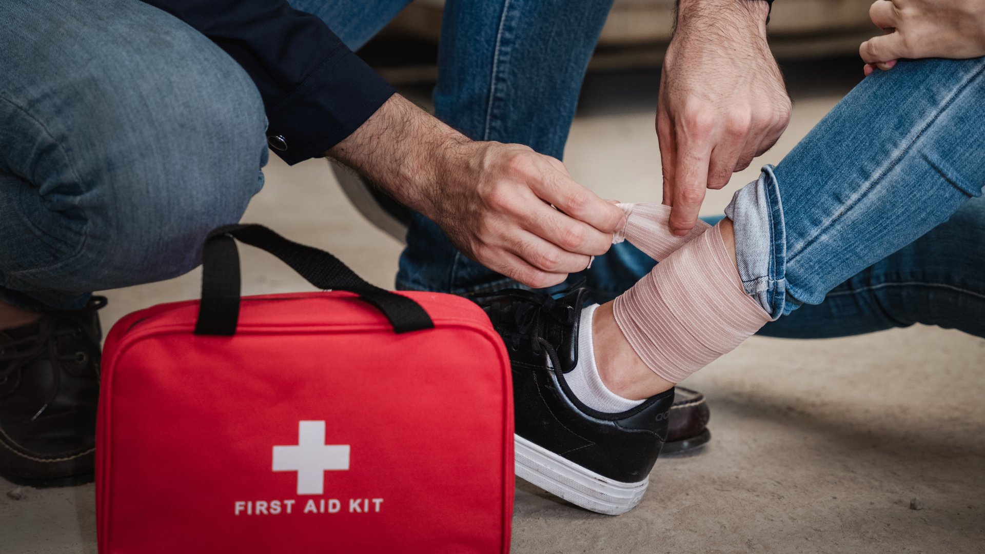 A worker administering first aid to a coworker. He is wrapping the coworker’s ankle with a bandage from the first aid kit.