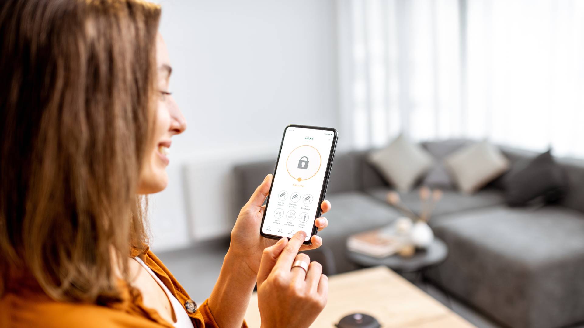 A young woman standing in her home, focused on her smartphone displaying a security application interface.