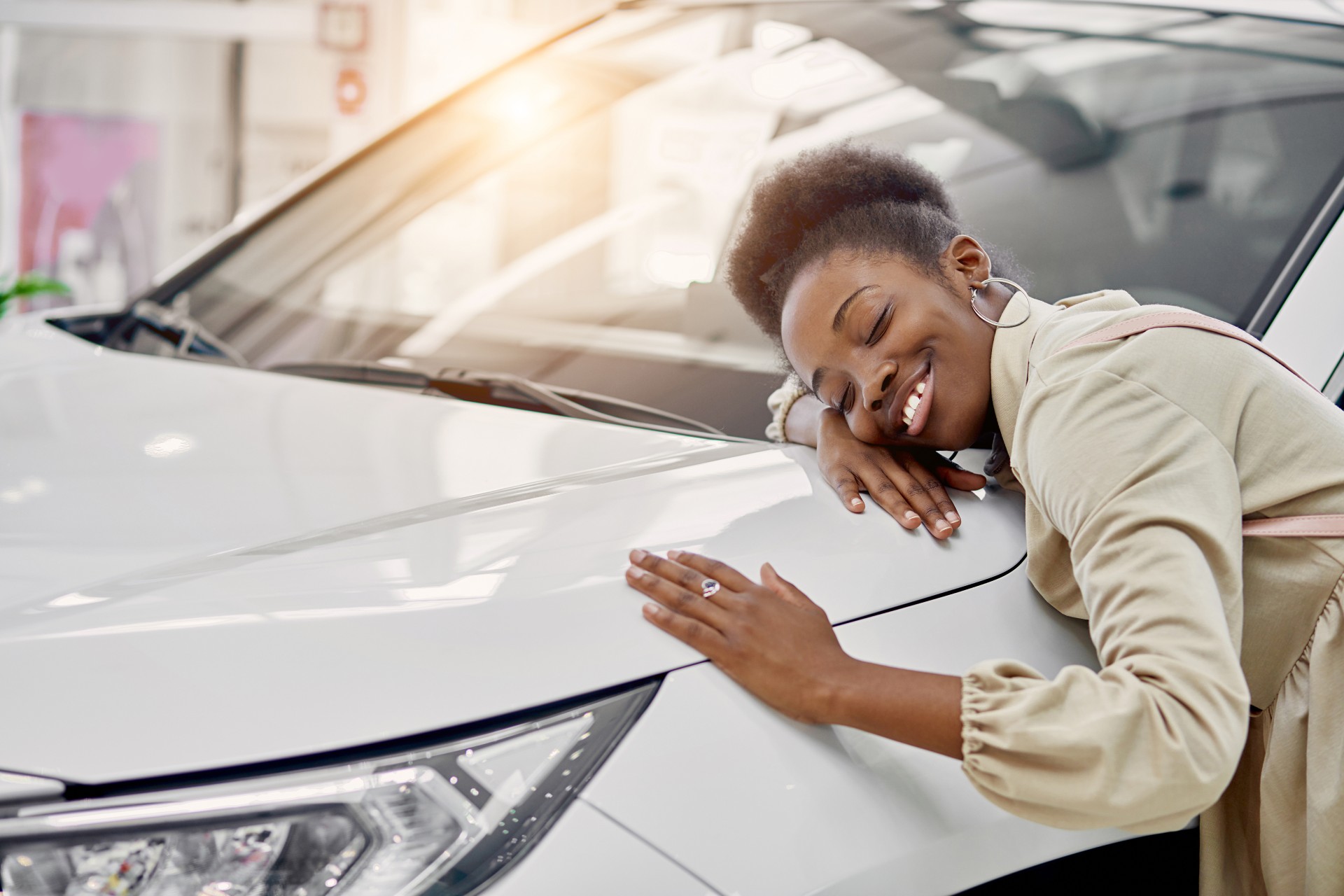 A Black woman wearing a beige dress smiles while touching a white vehicle with the sunlight reflecting off it.