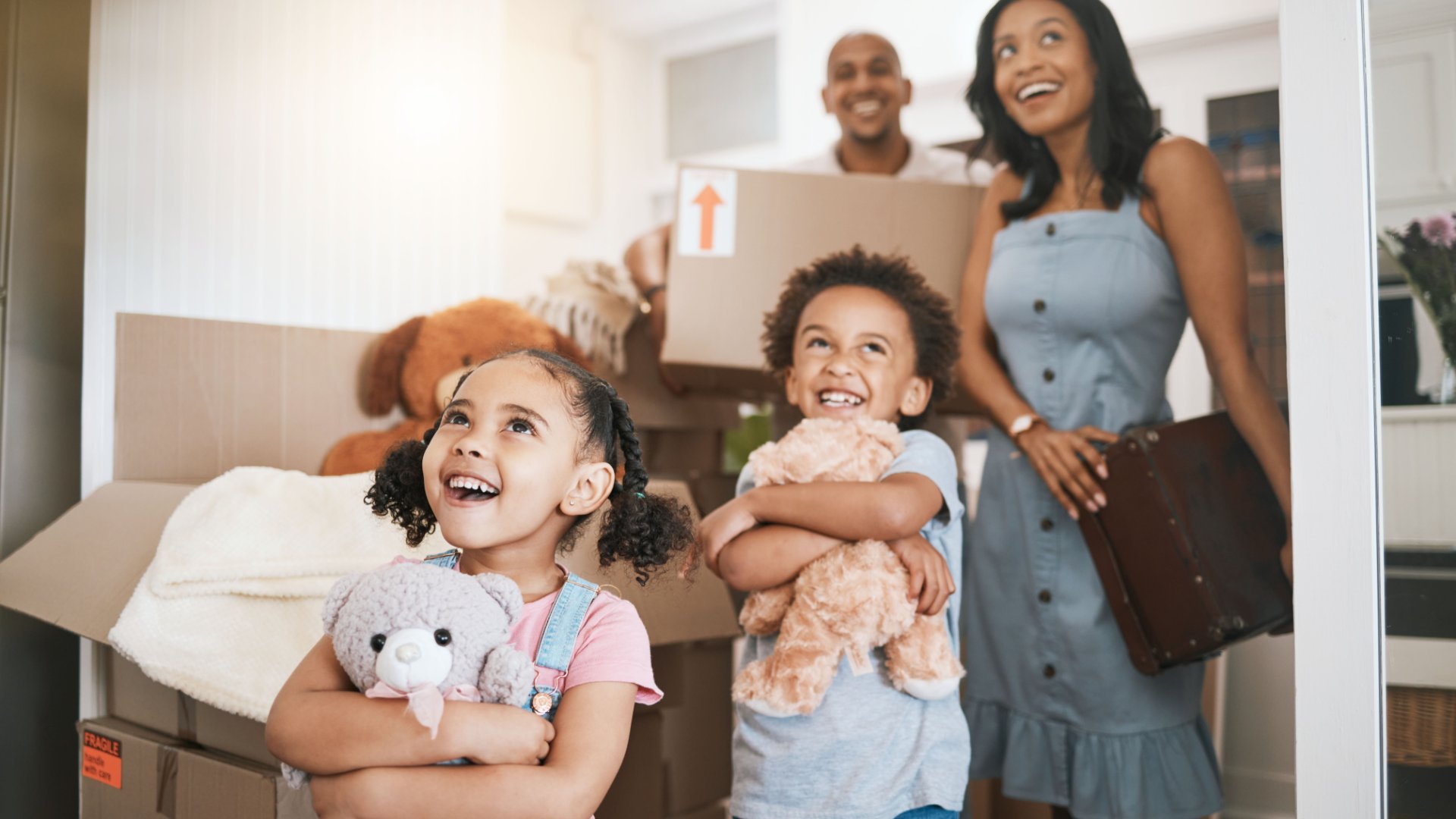 A family of four smiles as they enter a new home they have moved into. The two children both hold stuffed animals.
