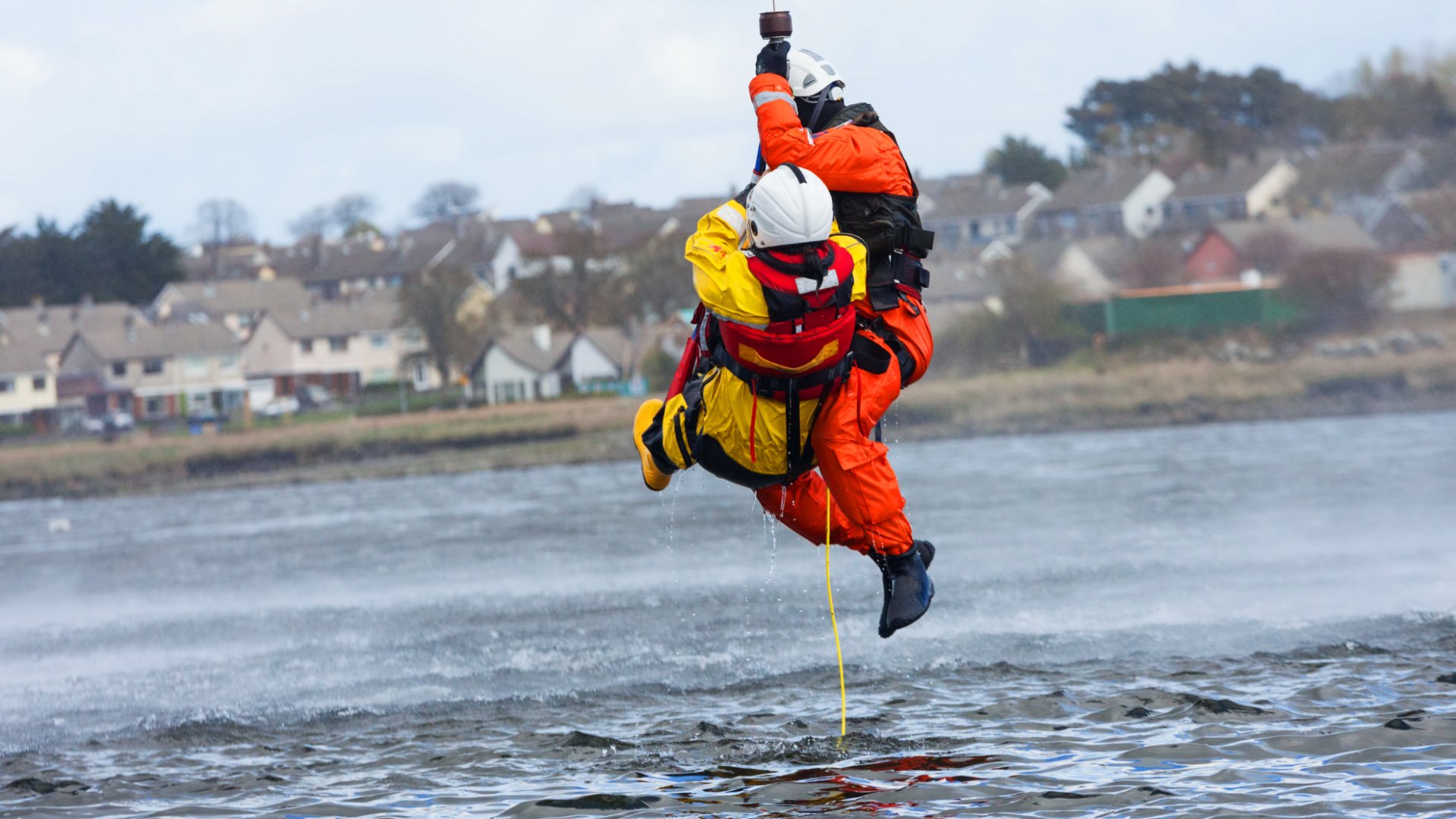 Two people wearing white hard hats and protective suits are being pulled from the water by a yellow rope.
