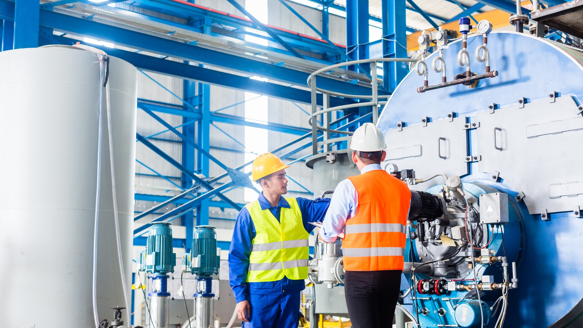 An employee and a manager in an industrial factory are talking to one another in the warehouse next to a large machine.