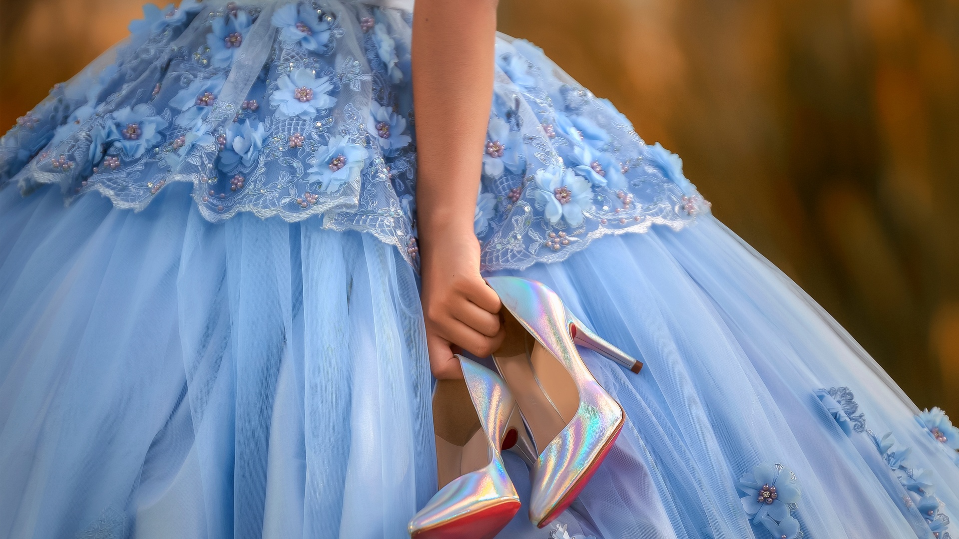 A close-up of a light blue quinceañera dress with lace and flowers. The young woman is holding a pair of blue high heels.