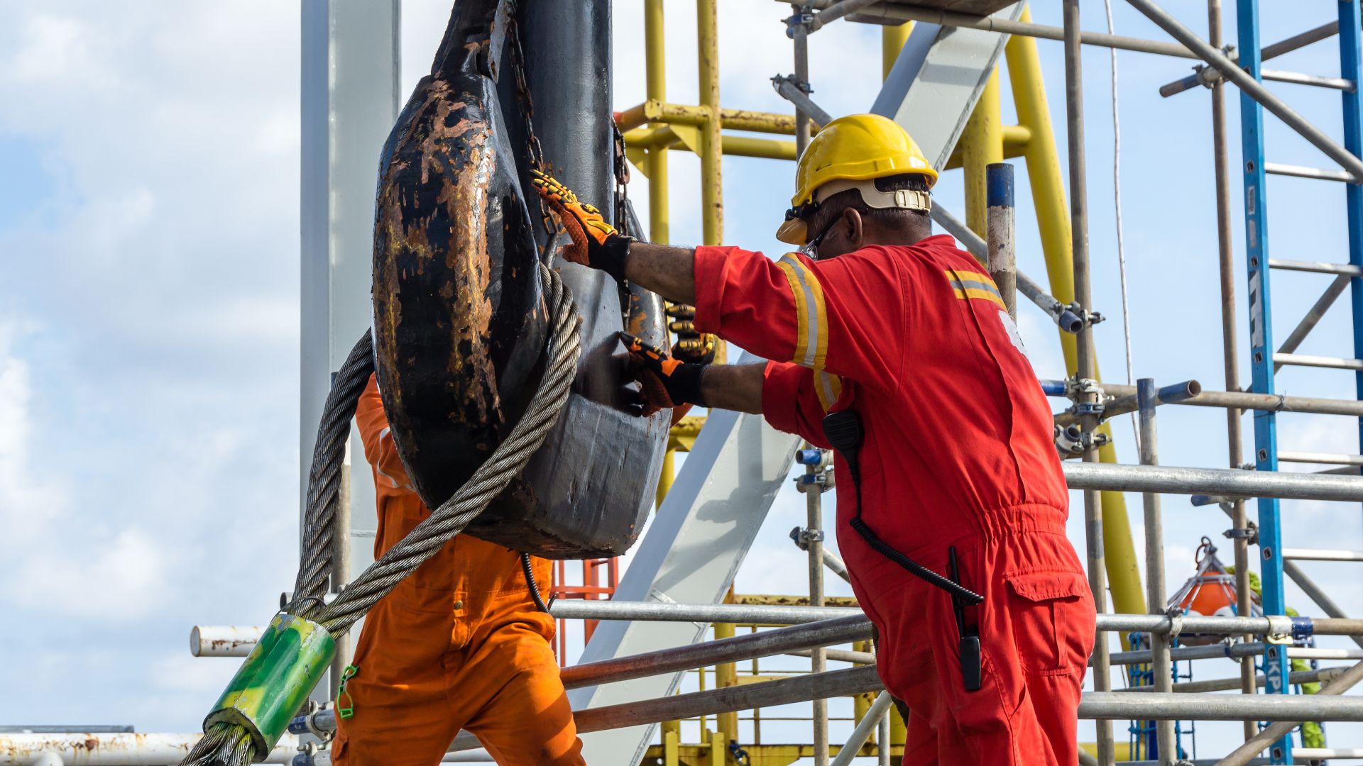 Two workers wearing orange and red jumpsuits attach a wire rope loop to a giant crane hook on a job site.