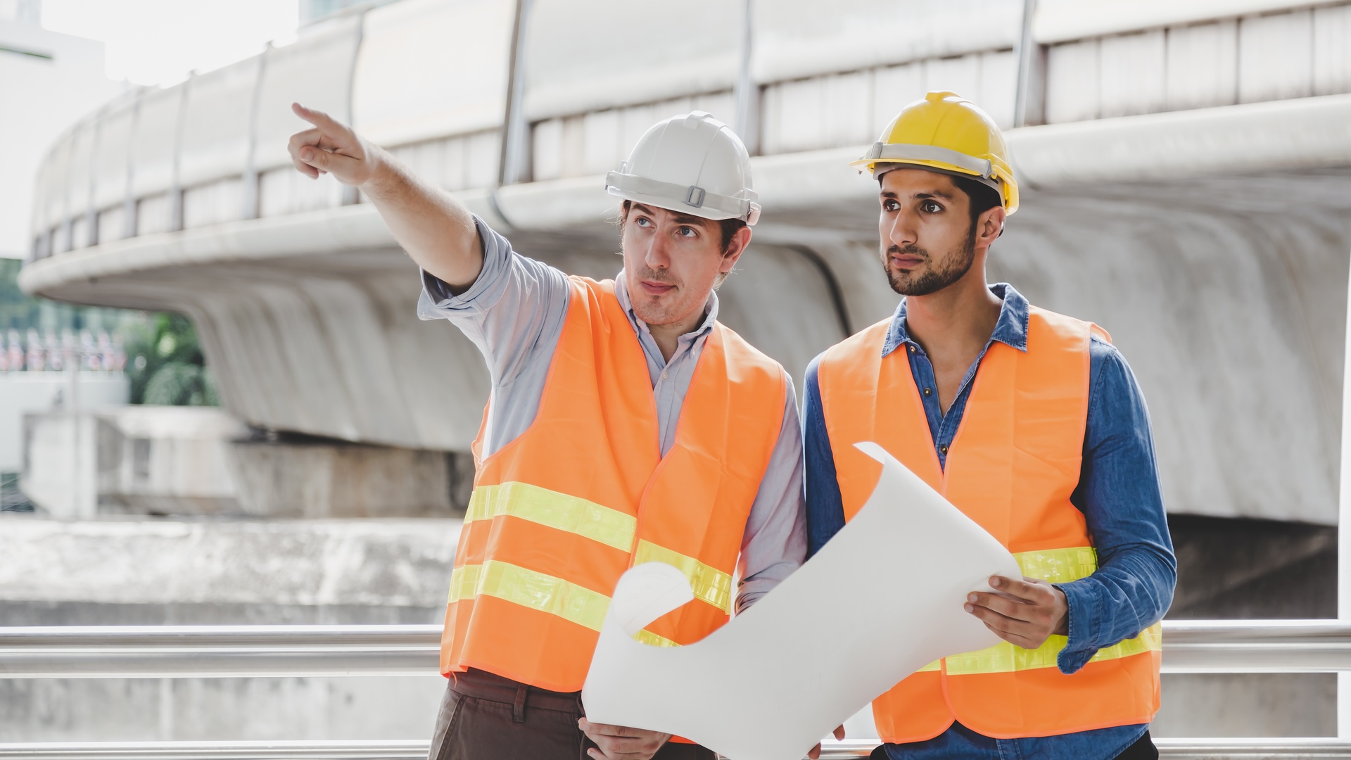 Two construction workers, wearing safety vests and white helmets, holding a large paper sheet, while looking ahead.