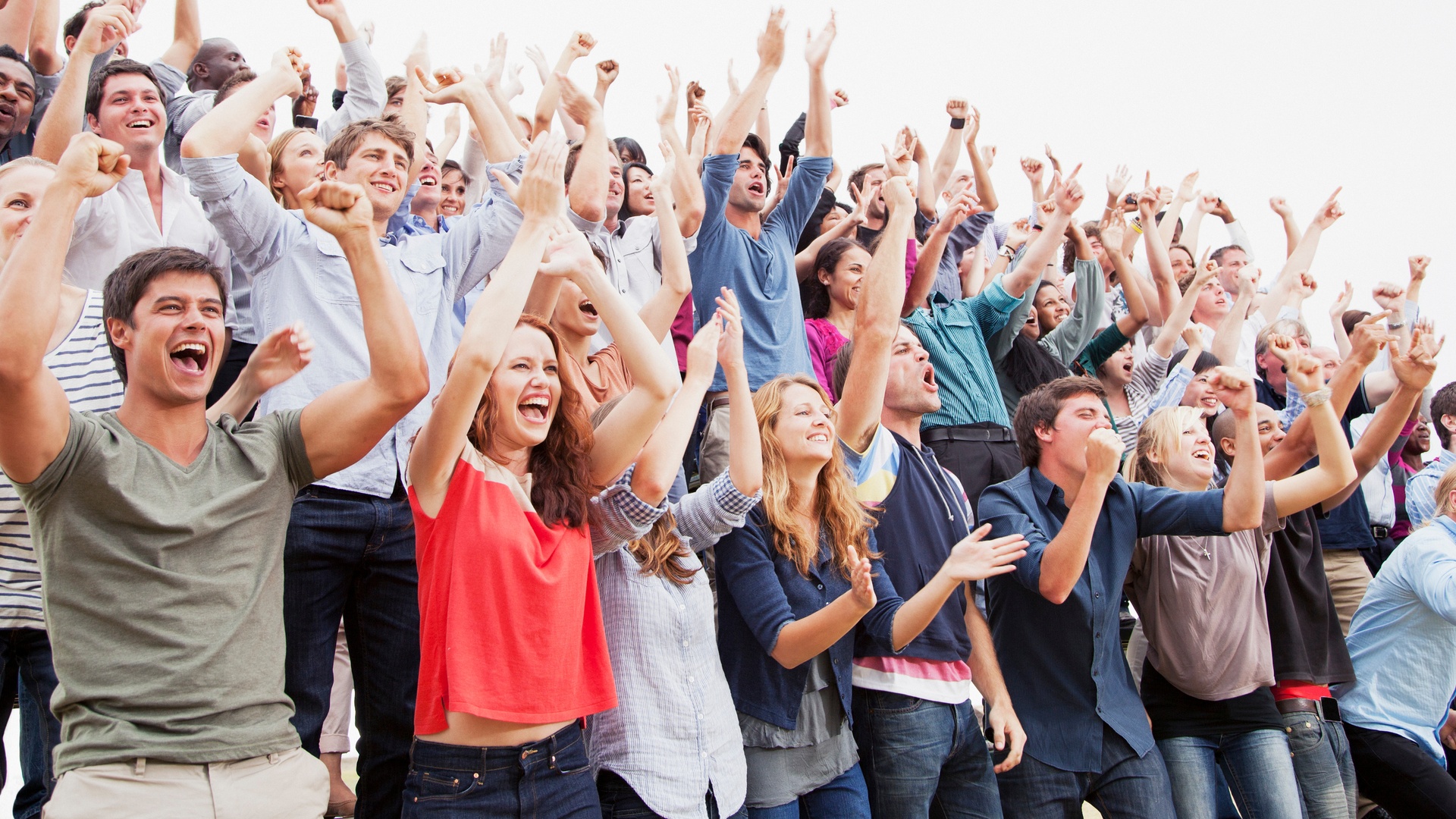 A diverse, cheering crowd with raised arms looks up in excitement, their joyful expressions lit by bright, even lighting.