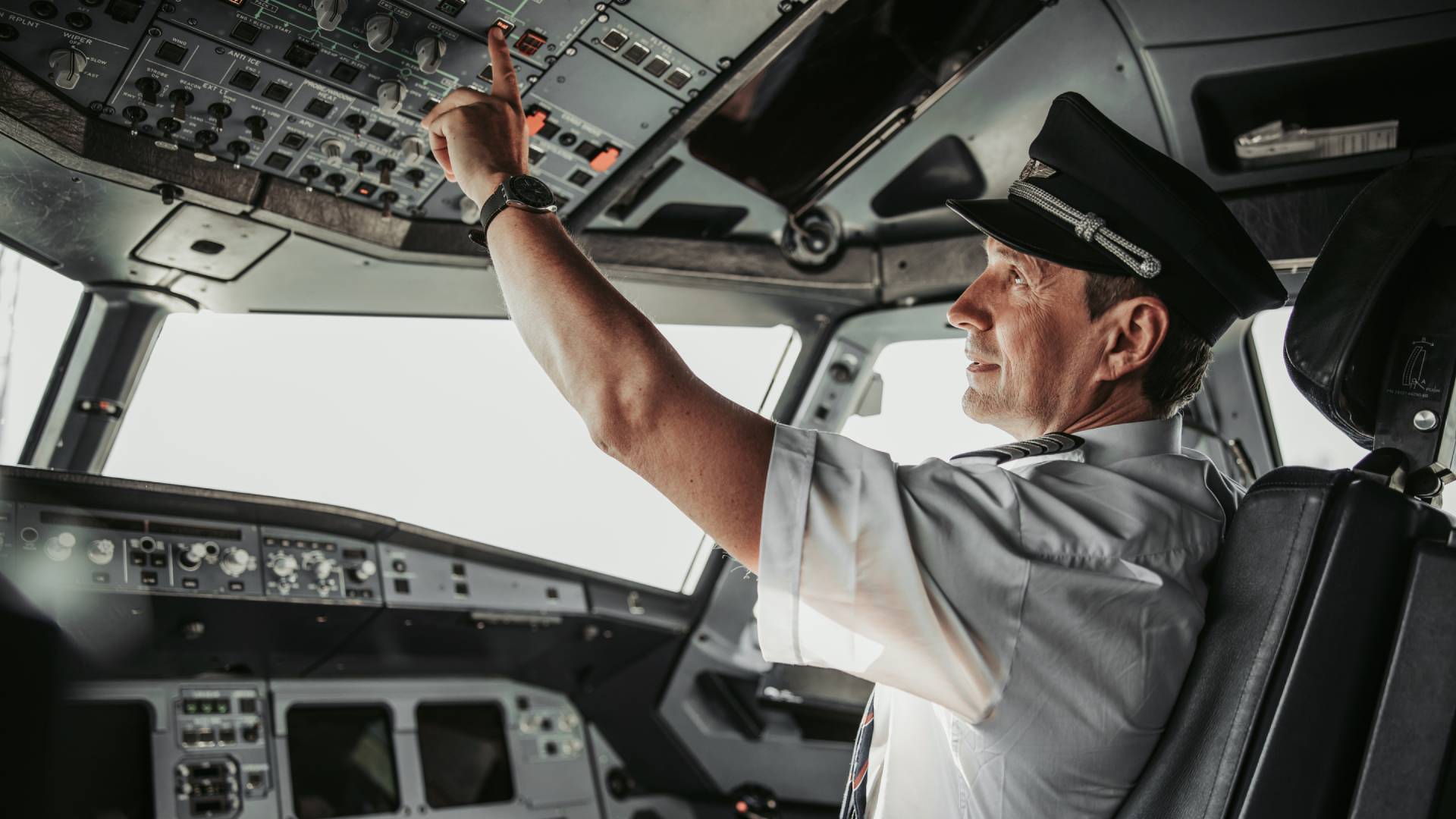 A uniformed pilot is sitting in the cockpit of an airplane. He is pushing a button on the ceiling of the plane.