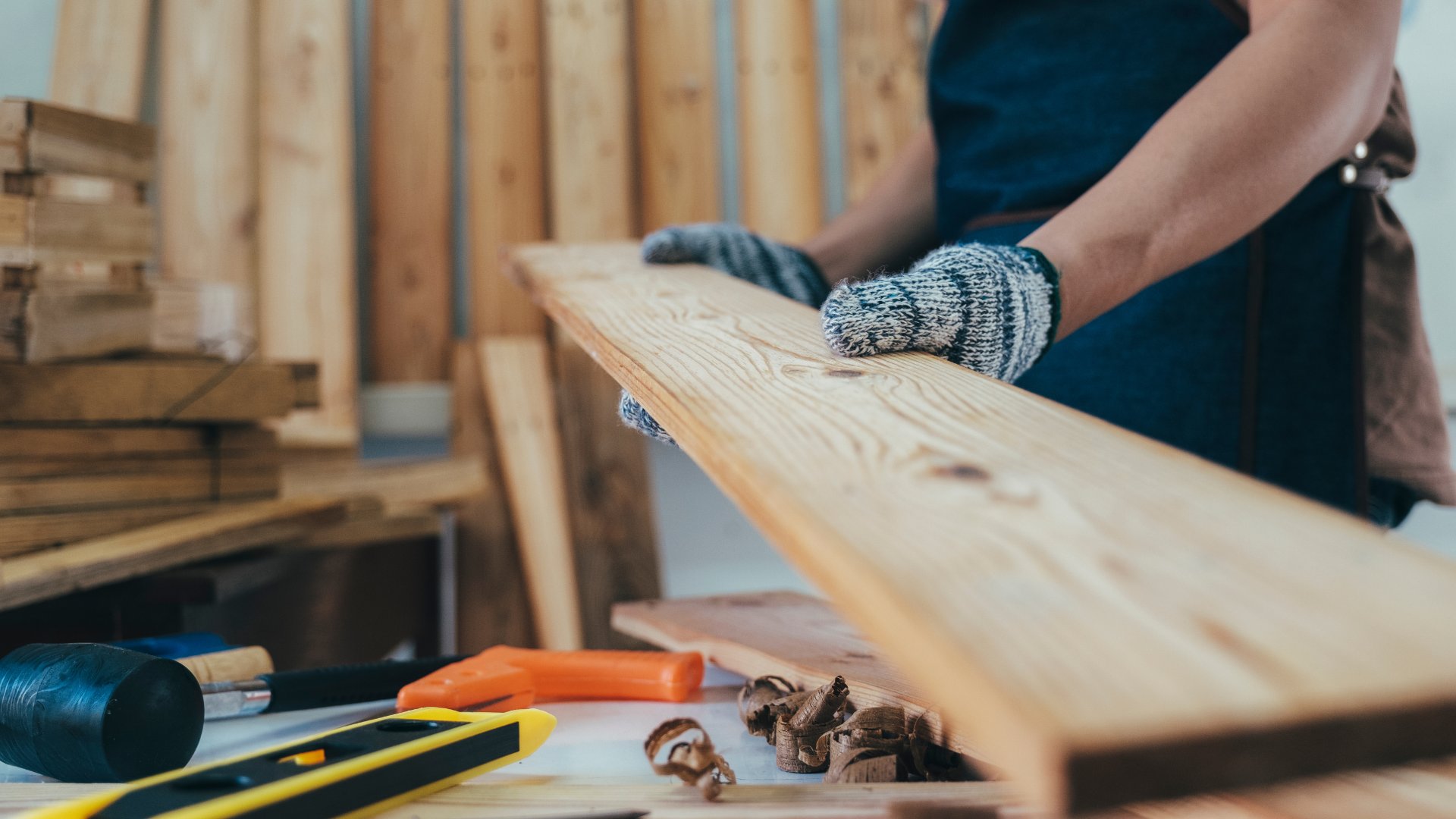 A woman in a workshop wearing safety gloves while handling a plank of wood. Tools are scattered on the table near her.