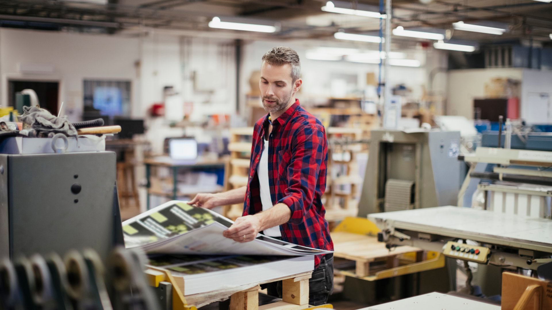 A man wearing a black and red plaid shirt is inspecting a large print that just came off an industrial printer.