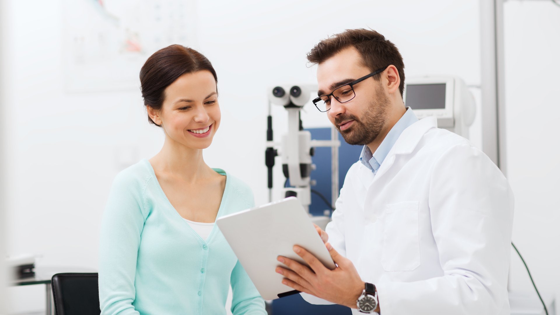 An eye doctor explains something to a happy patient, who looks at a tablet device in the doctor's hand.