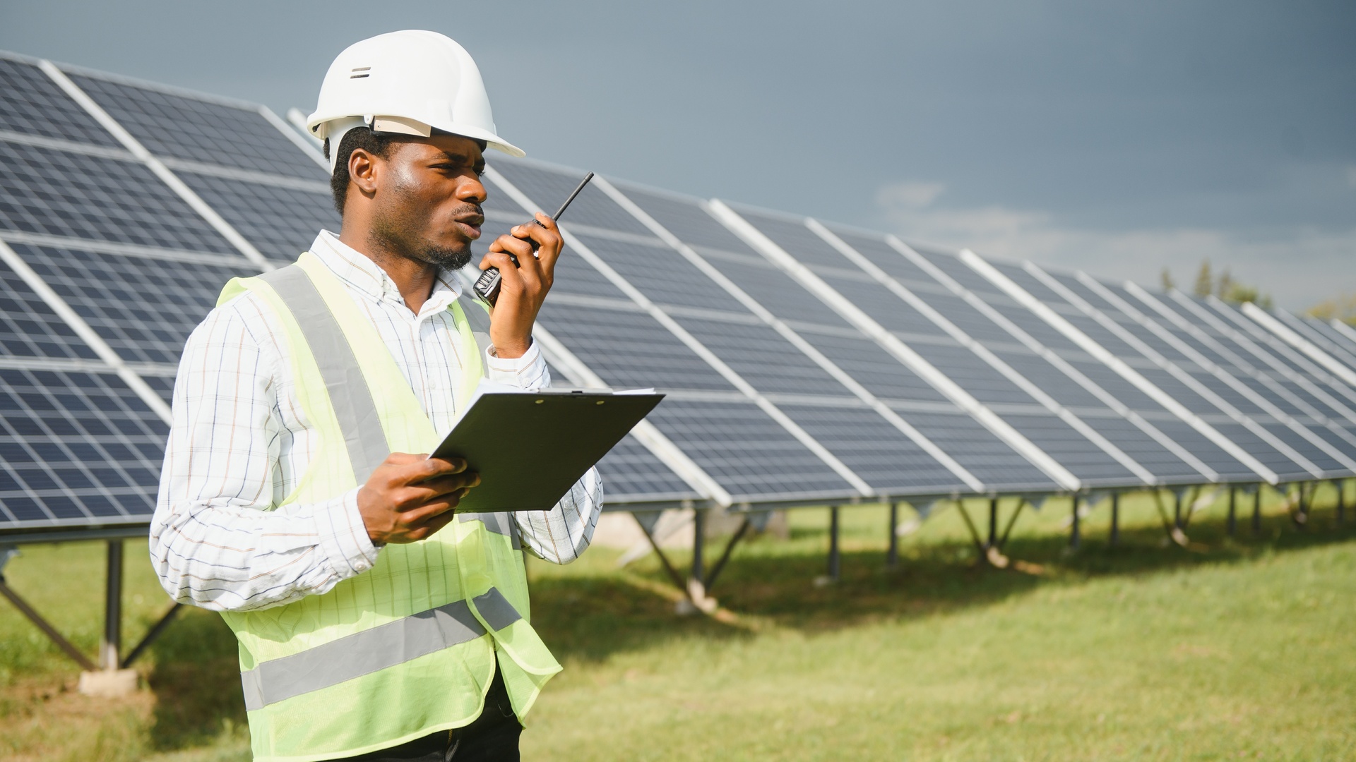 A man in a safety vest and hard hat using a two-way radio while standing in front of solar panels.