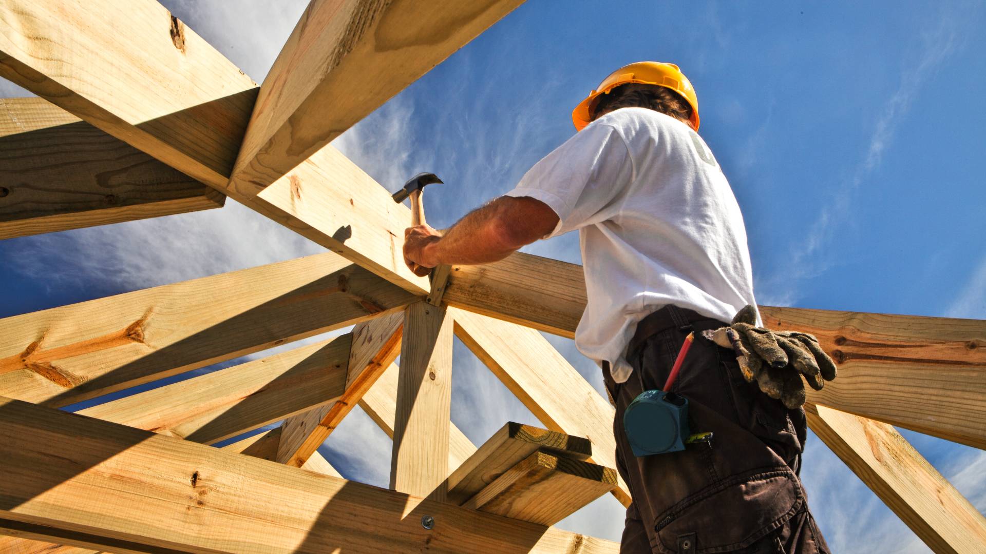 A man in a white shirt, brown cargo pants, and a yellow hard hat uses a hammer to nail a nail into a piece of wood.