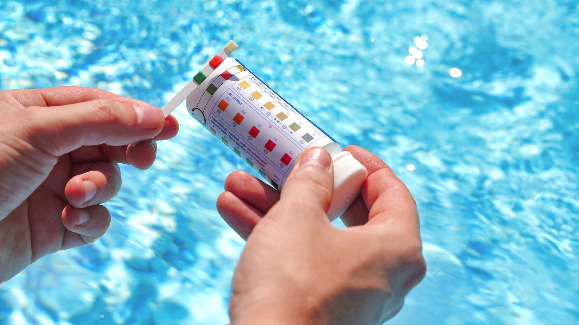 A close-up of a pair of hands using a pool water test with a test strip to determine PH value, chlorine, and algaecide.