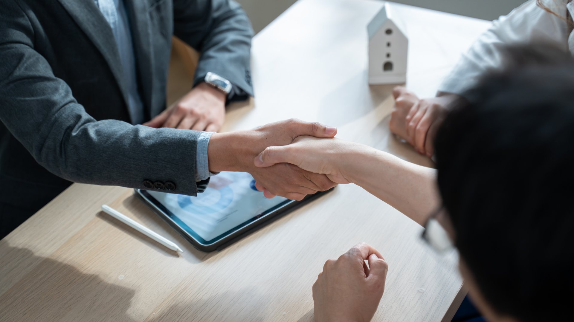 Two people shake hands across a desk that has a tablet, a pen, and a model house on it. A third person sits nearby.