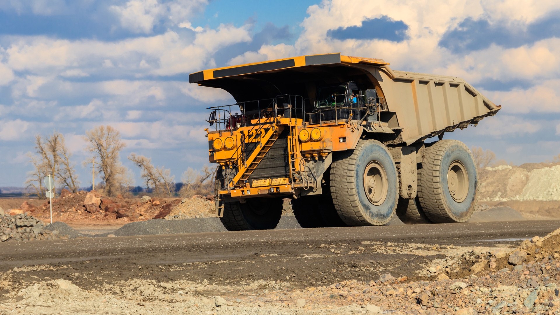 A dusty yellow haul truck with huge tires is parked on asphalt, surrounded by rocky desert terrain under a cloudy blue sky.