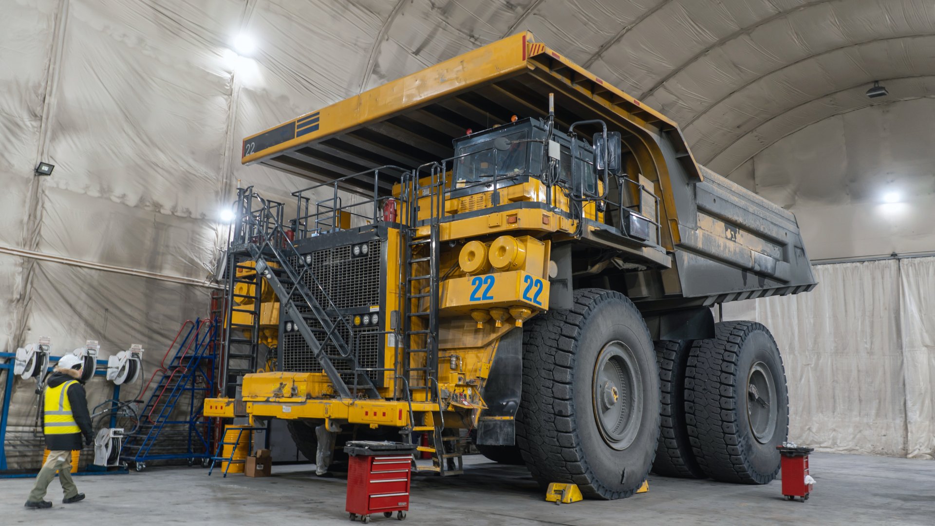 A large, yellow and black quarry dump truck in a large garage with a tool station next to it and a worker approaching.