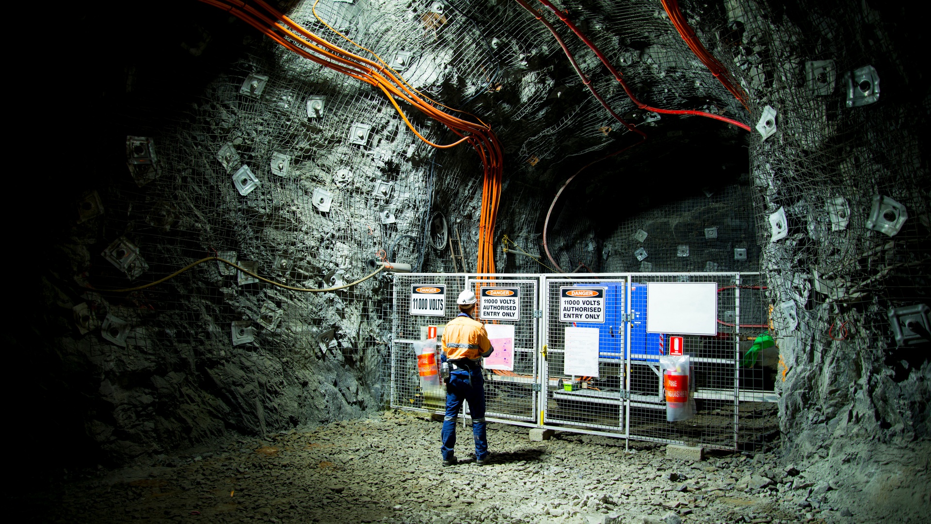 A worker wearing an orange vest stands in an underground mine. A fence with a warning sign blocks electrical components.