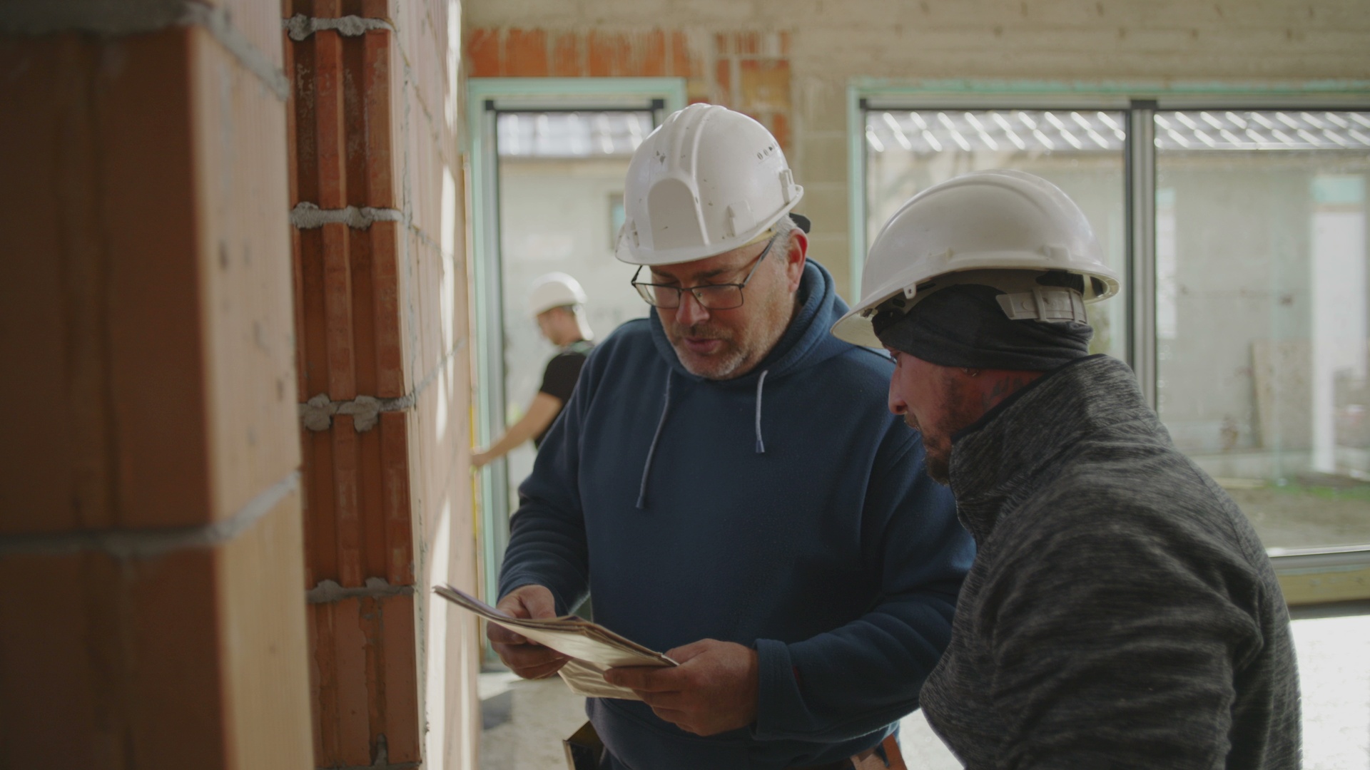 Two construction workers are measuring a brick wall at a construction site with exposed walls and large windows.