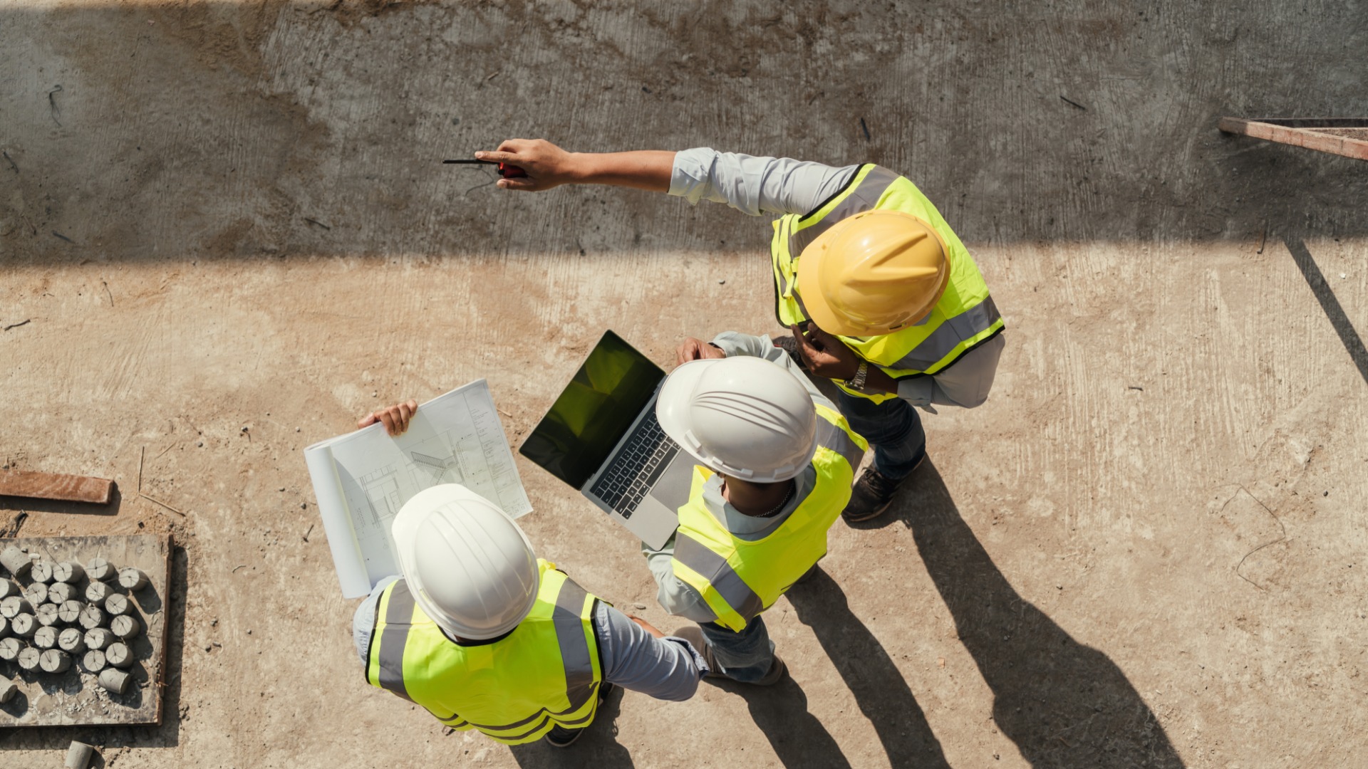 Three men in hard hats are standing next to each other. One man is holding a blueprint and another is pointing.