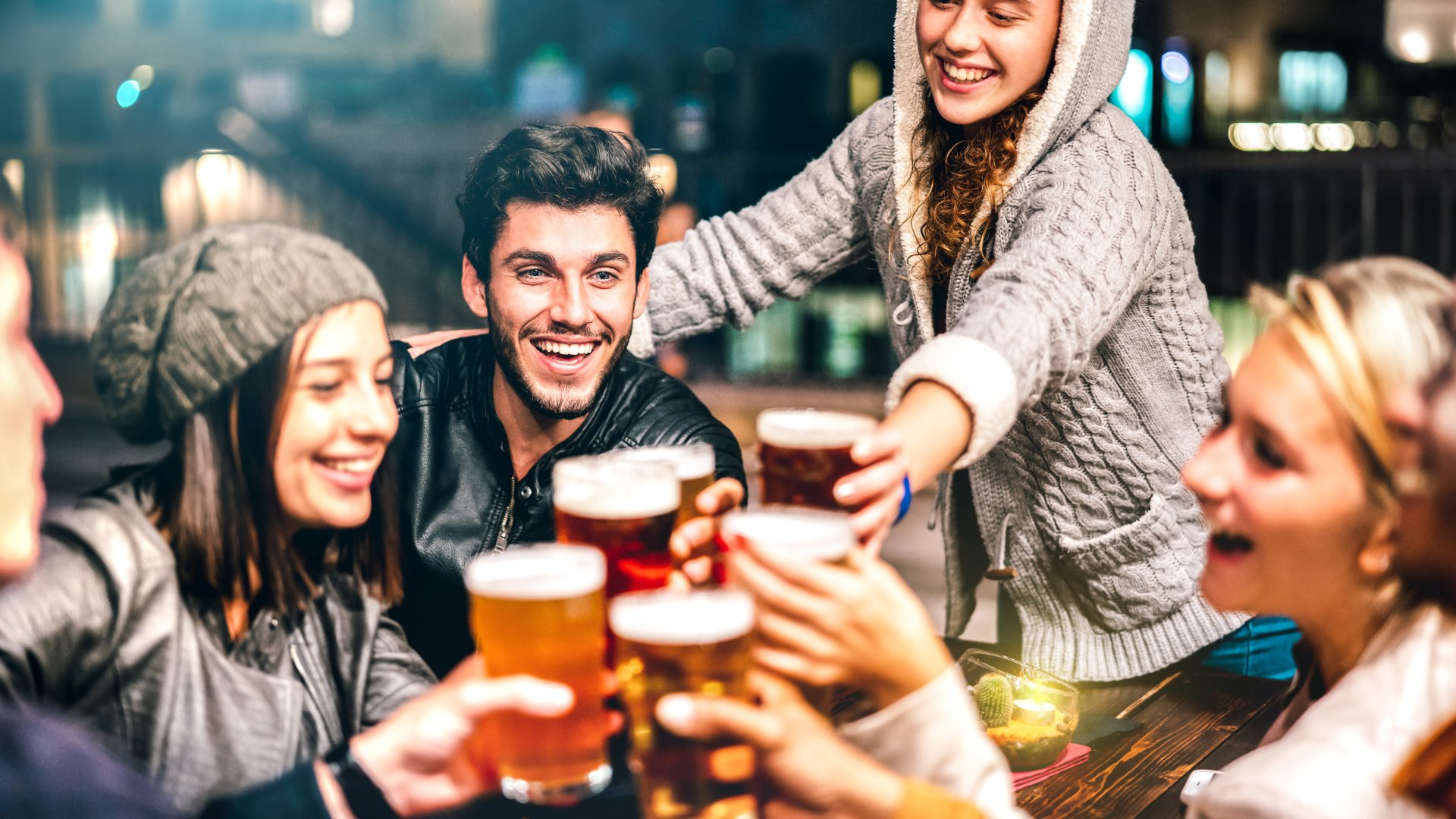 A group of six friends in winter clothing cheers with their beers over a table at an outdoor bar.