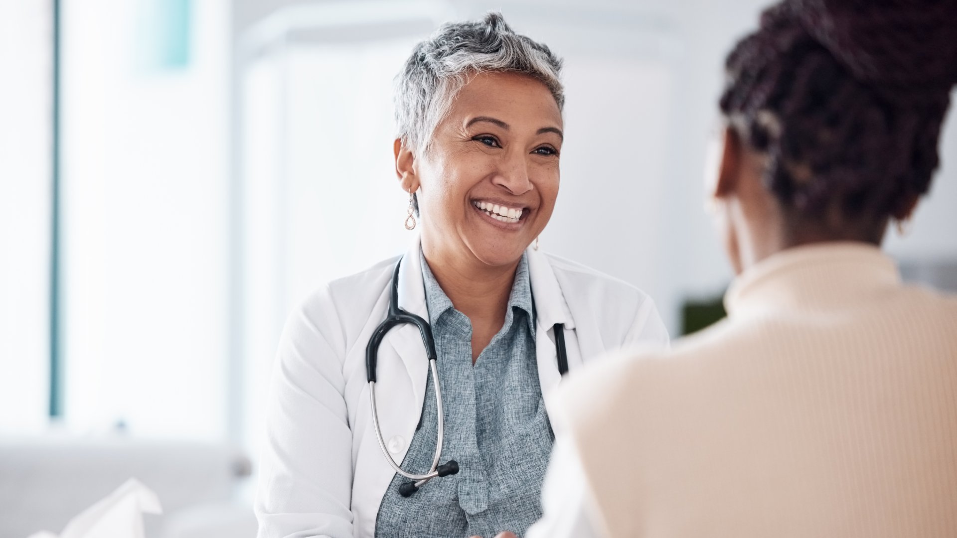 An older medical professional smiling at her patient. She's wearing a white coat and has a stethoscope around her neck.