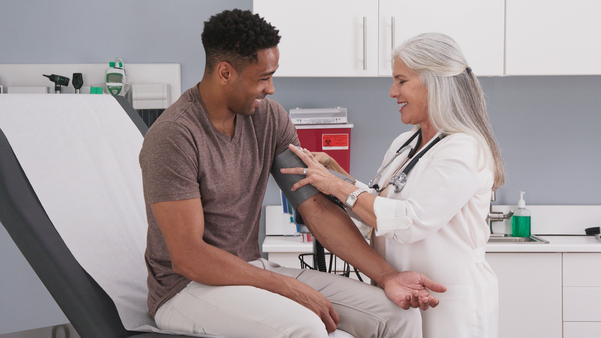 A man is sitting on an exam room table. There is a woman in a white coat grabbing the man’s arm.