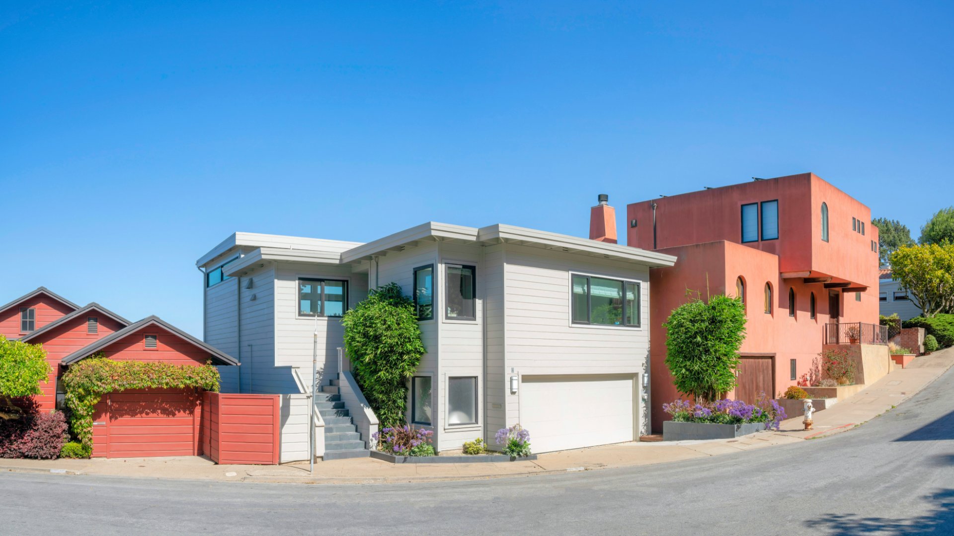 A two-story white house between two orange houses on a sloped concrete street with some trees on a sunny day.