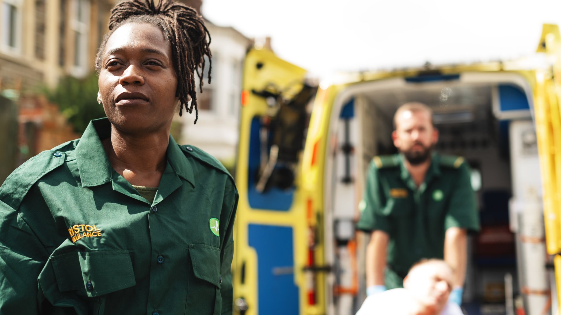 A man and a woman are transporting a person on a gurney. A yellow emergency vehicle is behind them.