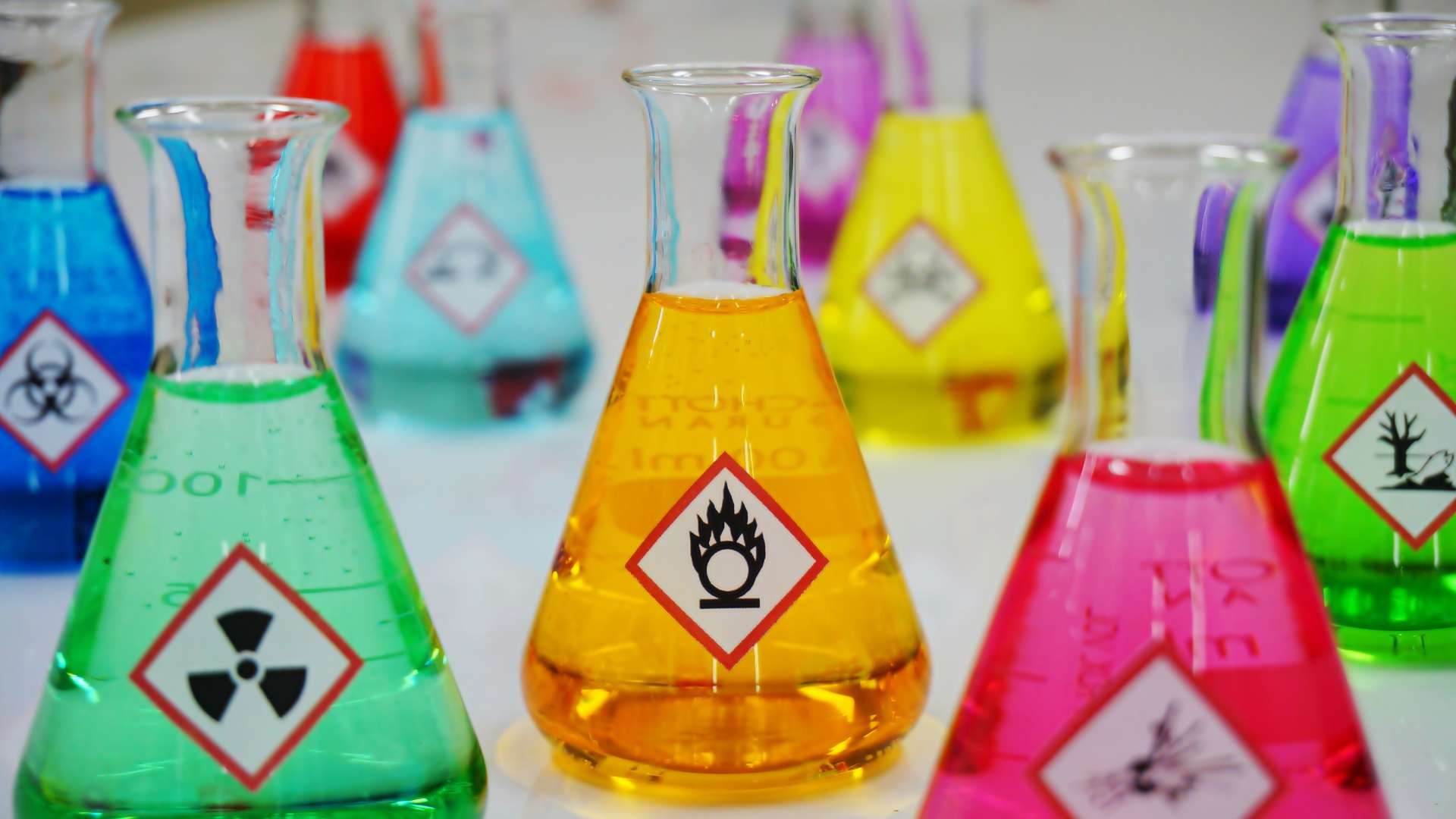 Multiple Erlenmeyer flasks filled with colorful chemicals. Each flask has a different hazardous warning symbol on the front.