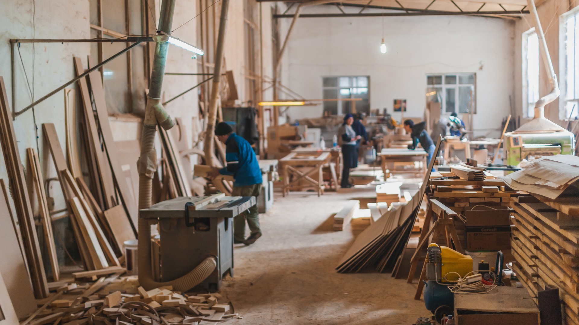 Busy carpentry workshop with workers shaping wood, sawdust on floor, and planks stacked along walls.