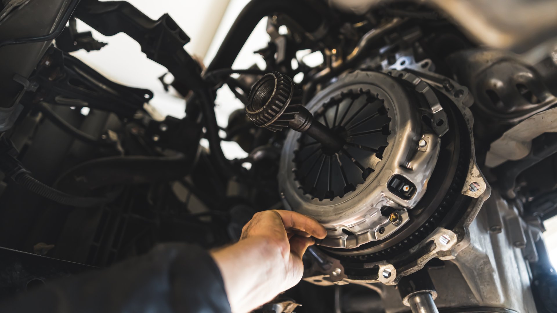 A close-up of a mechanic's hand reaching into the undercarriage of a lifted vehicle to touch the exposed clutch.