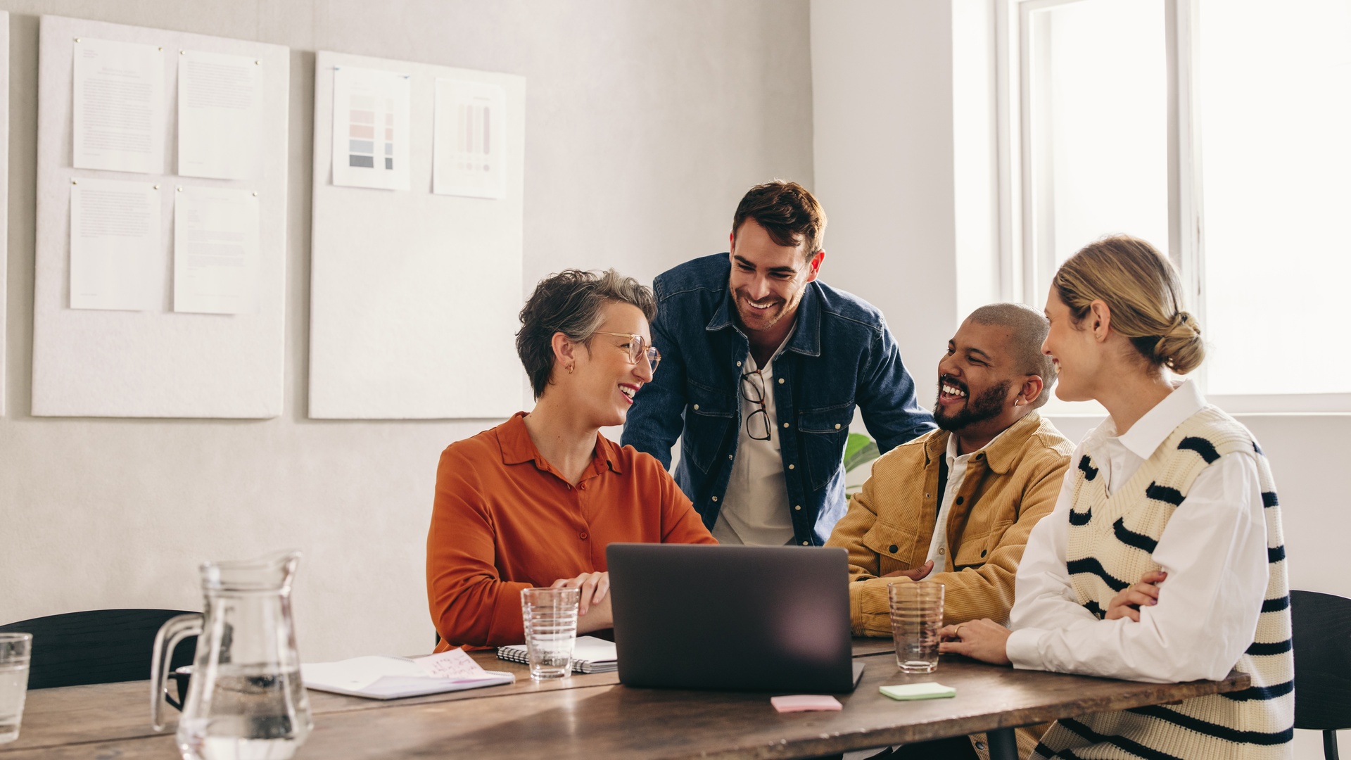 A group of four smiling coworkers having a meeting in a conference room with white bulletin boards on the wall.