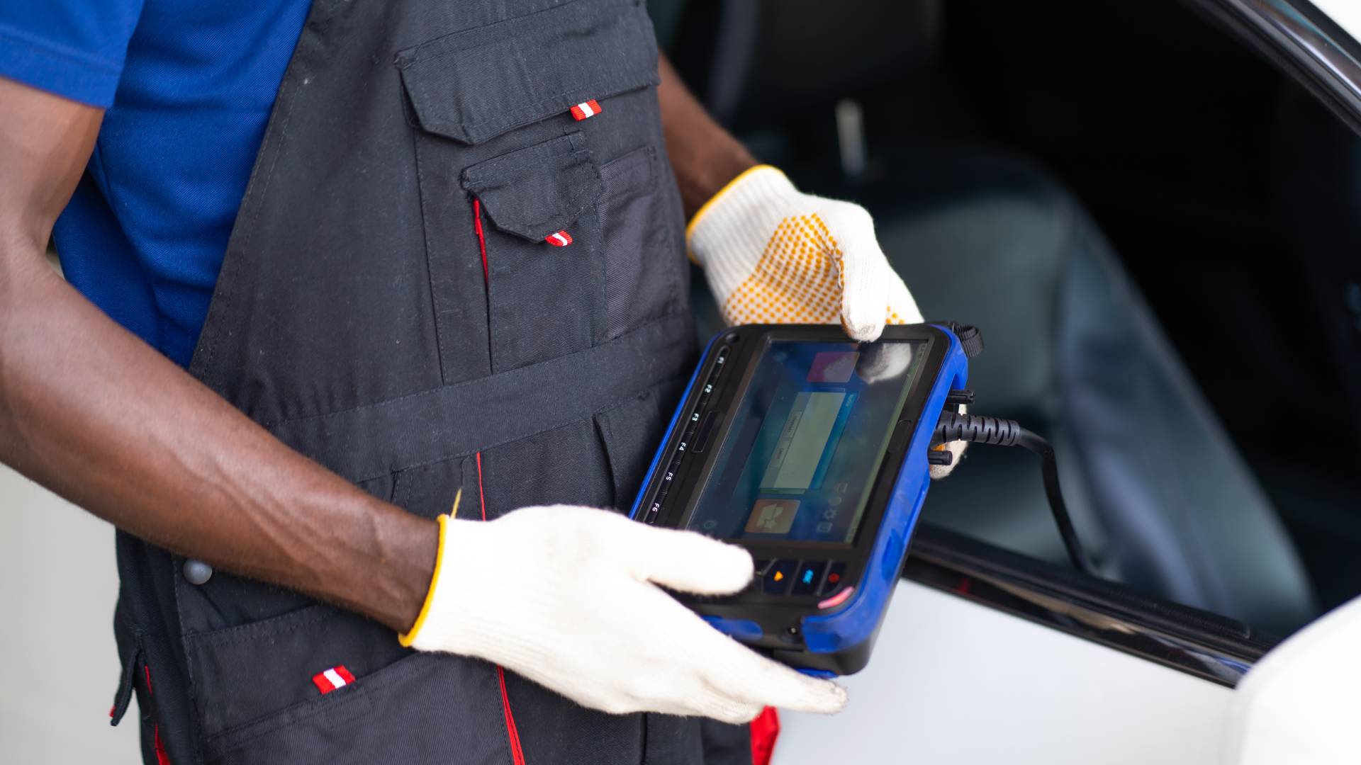 A mechanic in overalls and gloves is holding a device with a screen to read data from a car's engine control unit.