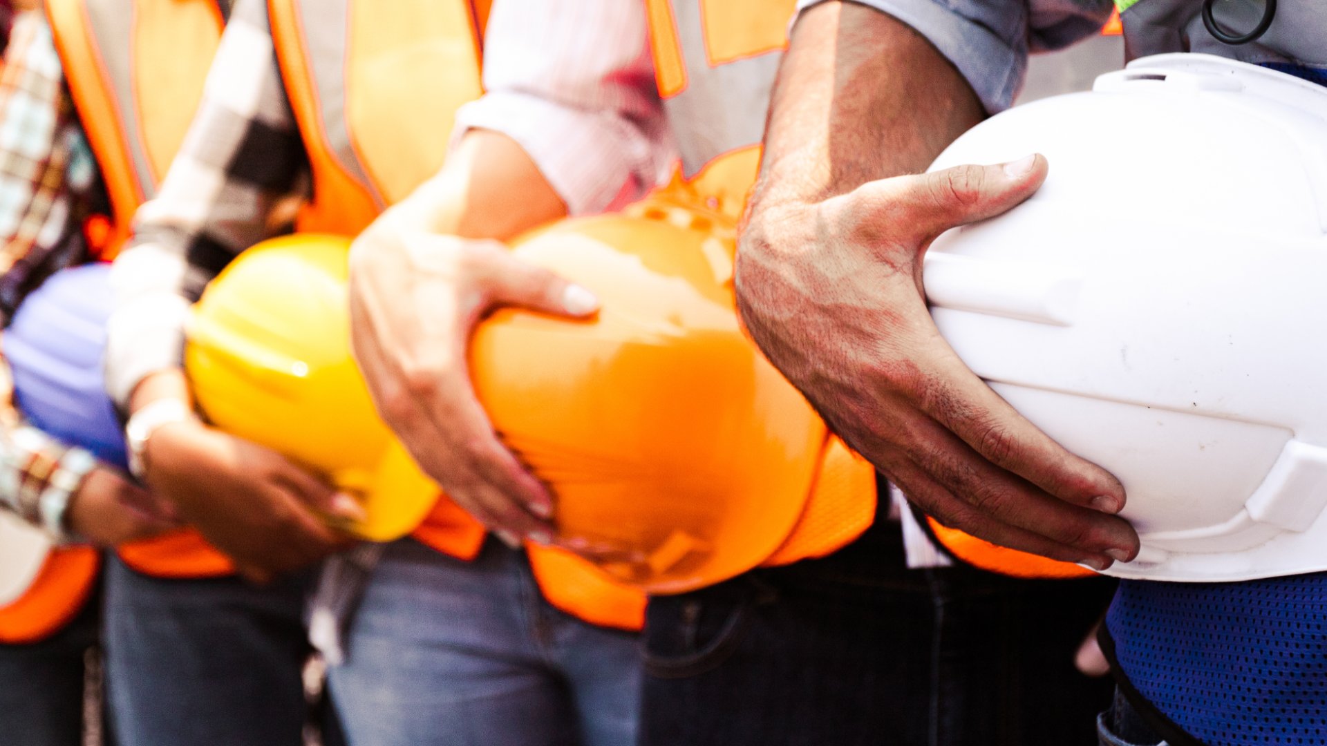 A close-up of a group of workers in a line, all wearing high-visibility safety vests and holding hard hats.