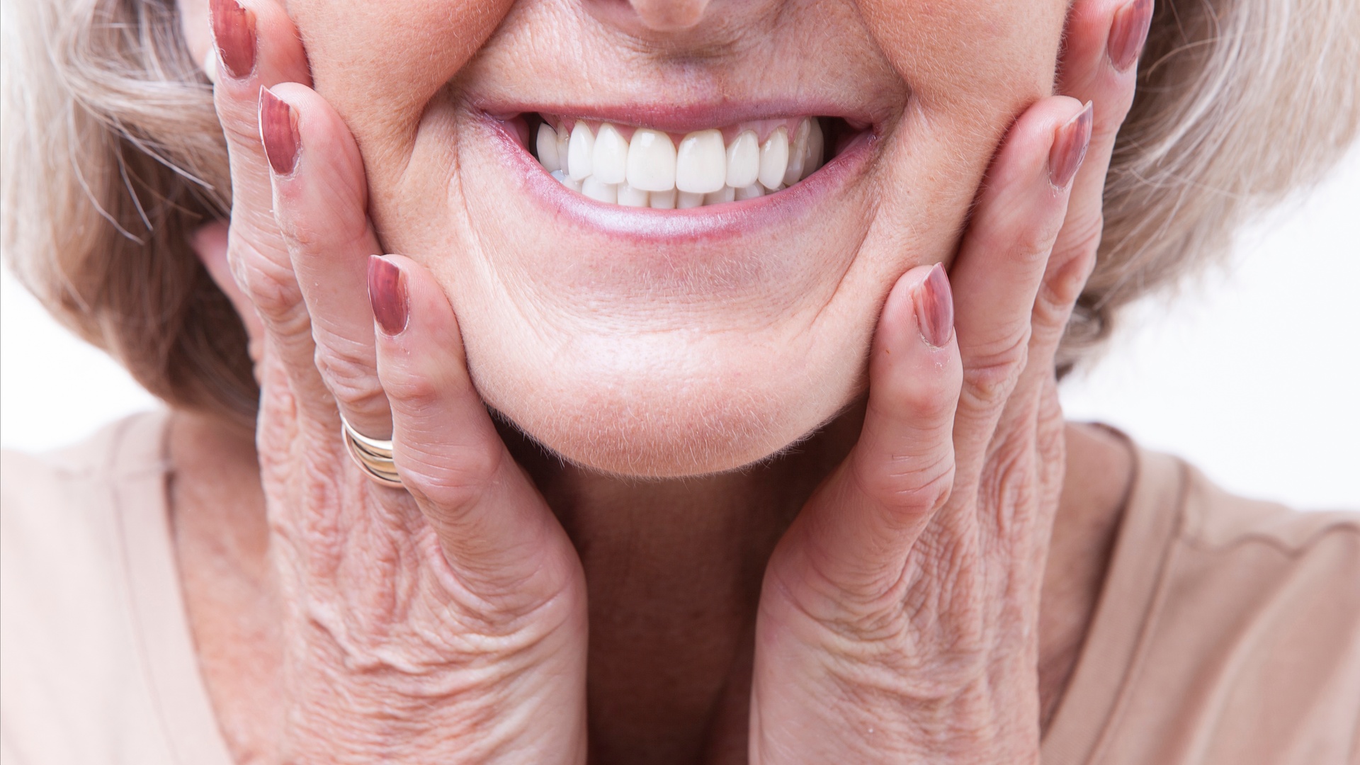 A close-up of a smiling senior woman with her hands on both sides of her mouth showing off her white teeth.