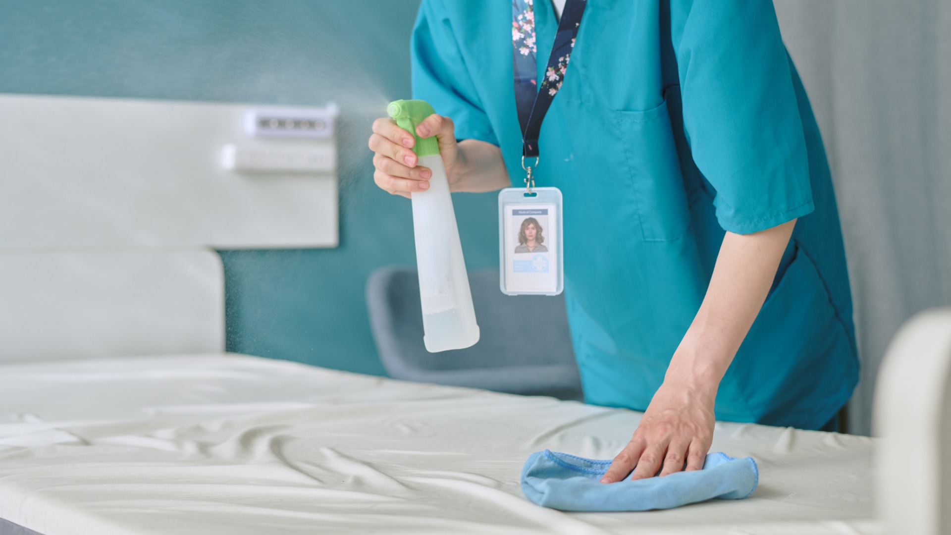 A woman wearing a blue uniform and a lanyard with a credential while disinfecting the top of a bed mattress.