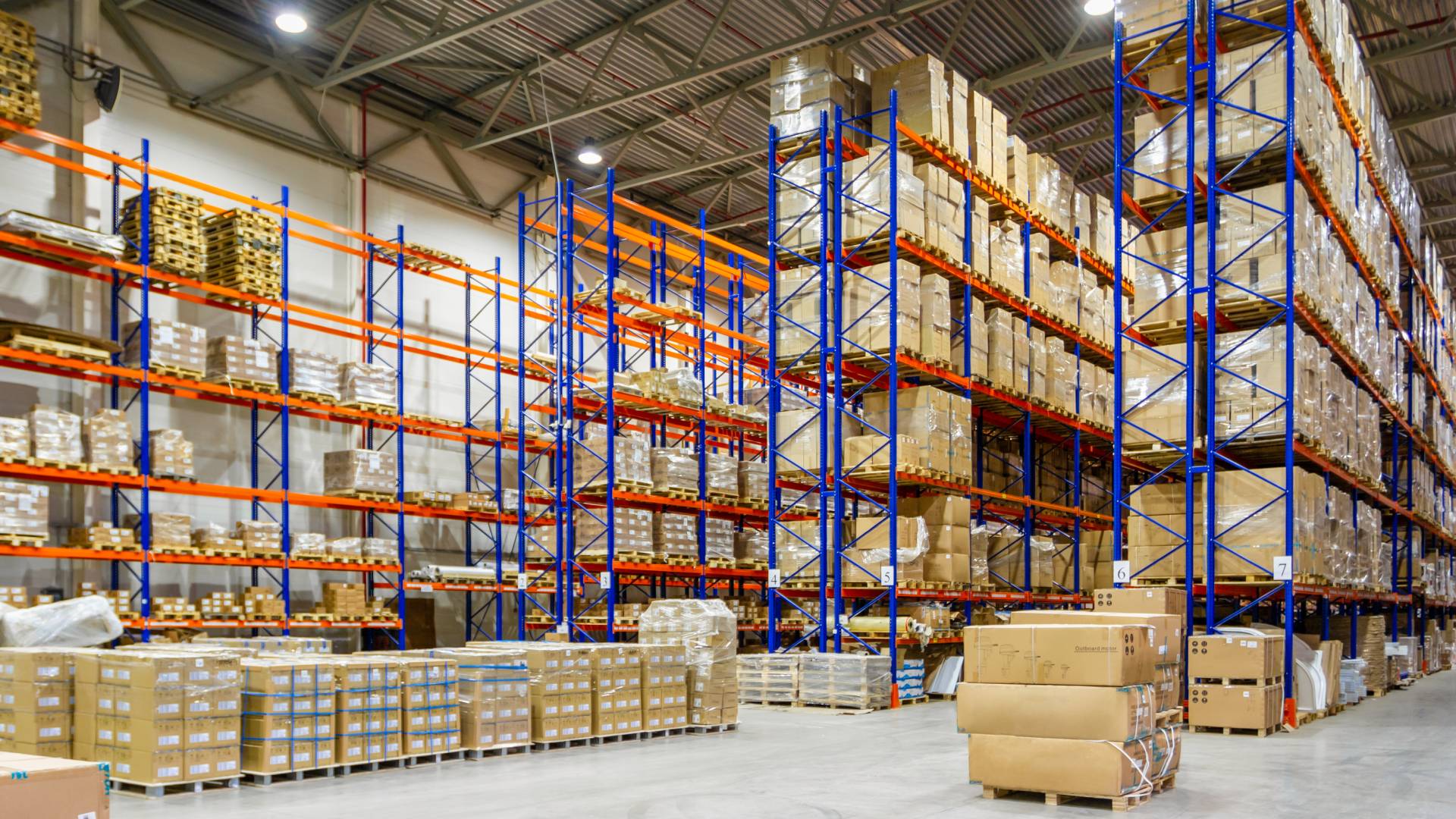 The interior of a large warehouse with an extensive pallet racking system. The pallets are filled with brown cardboard boxes.